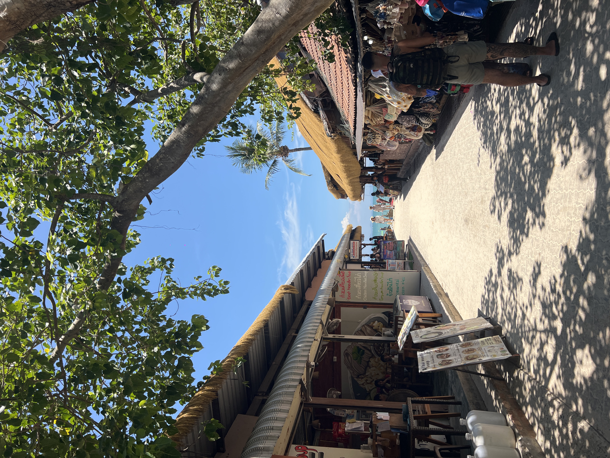 The image depicts a street scene in Ban Ao Nang, Thailand. A concrete walkway extends towards a beach with visible blue water and sky. Along the walkway are several businesses with thatched roofs and signs, including a restaurant advertising Thai noodle soup and chicken dishes. People are visible in the distance, some appearing to be tourists. In the foreground, there is a tree with leafy branches, casting shadows on the walkway. There are also several menu boards displayed outside the restaurant. One person is visible along the walkway, looking at merchandise displayed at another business, and the person has tattoos on their legs.
