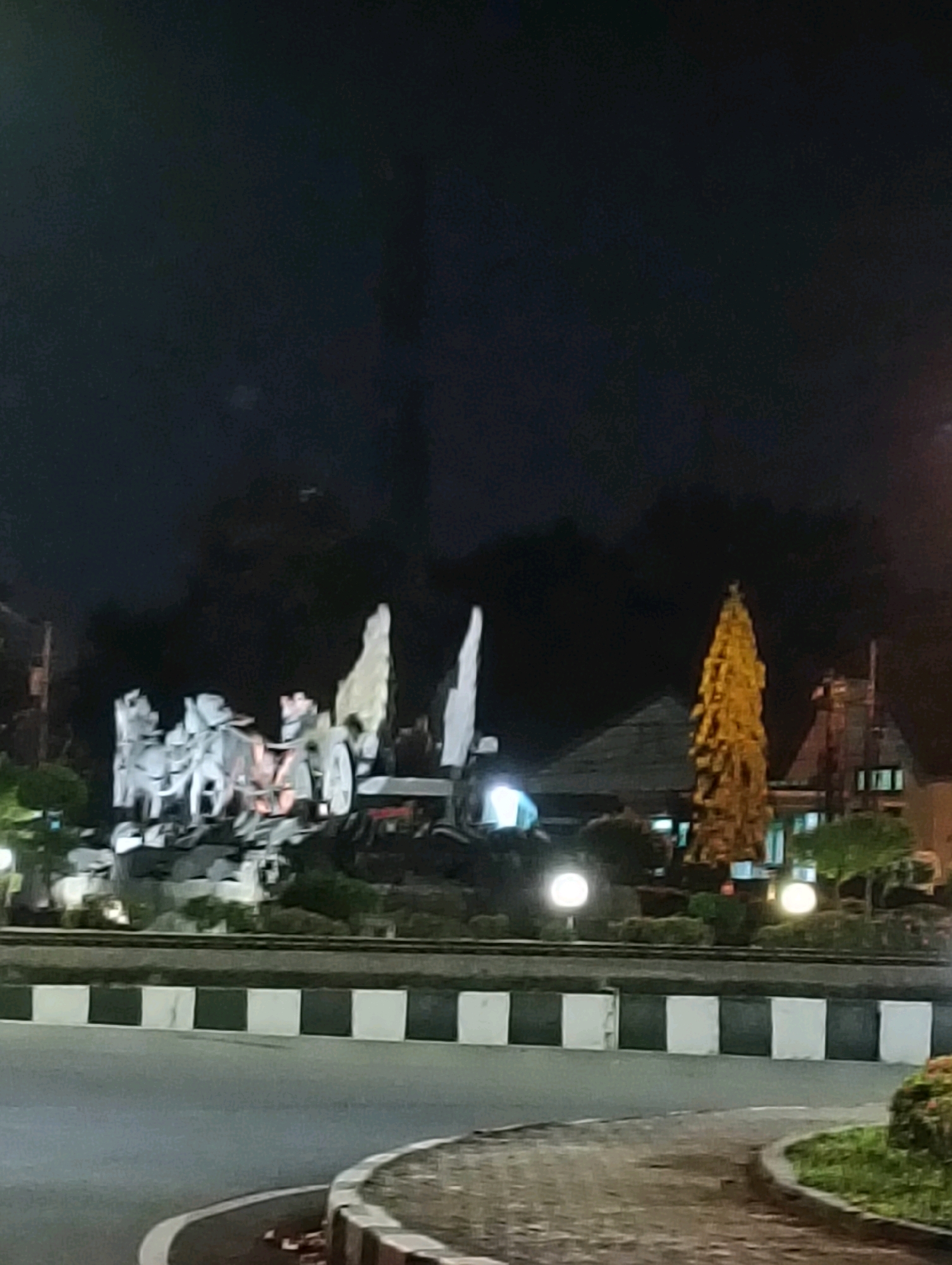 This is a nighttime photograph taken from a slightly elevated position, likely from a vehicle. The scene depicts a roundabout in the city of Purworejo, Indonesia. In the center of the roundabout, there is a prominent monument with several abstract, white sculptures. Behind the monument, a golden, conical tree-like structure stands tall, illuminated from within. Further in the background, buildings with lights are visible. The roundabout itself has a distinctive black and white checkered border along its edge, separating it from the asphalt road. Streetlights are also present, casting a soft glow on the surroundings. The overall impression is of a well-lit public space at night.