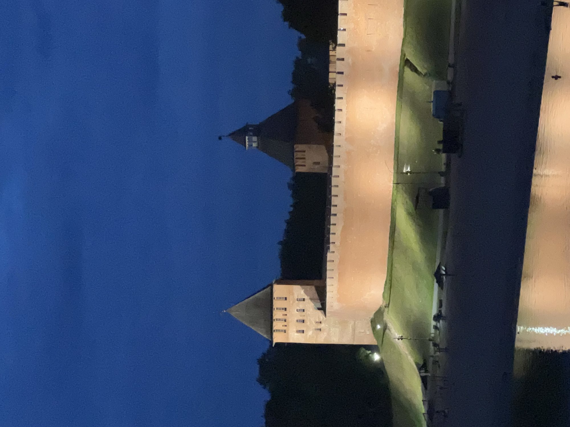 The image shows the Veliky Novgorod Kremlin at dusk. The imposing brick walls of the kremlin are illuminated by artificial lights, casting a warm glow on the ancient structure. Two prominent towers rise from the walls, their conical roofs silhouetted against the deep blue twilight sky. The kremlin is situated on a grassy embankment that slopes down towards a body of water, possibly the Volkhov River, which reflects the illuminated walls and sky. In the foreground, the dark water of the river is visible, with some faint reflections. The embankment is dotted with a few lampposts and some indistinct figures, likely people enjoying the evening. The overall atmosphere is serene and historic.