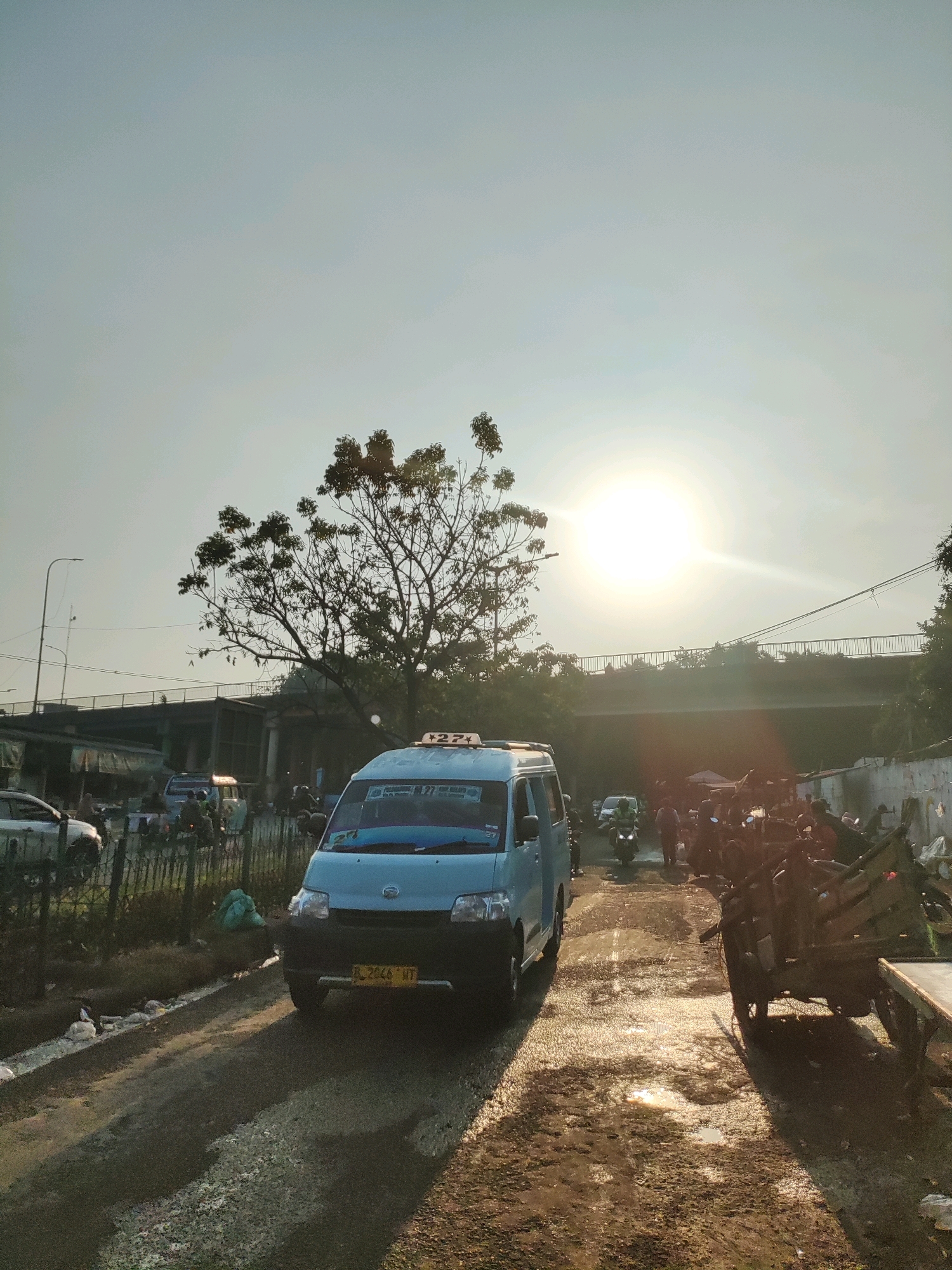 The image depicts a street scene in East Jakarta, Indonesia, during what appears to be late afternoon or early morning, given the low angle of the sun and its bright glare in the sky. A light blue van, possibly a taxi, is prominently positioned in the foreground, facing left. The van has a license plate that reads "B 2846 WT". On its roof is a sign with "27+" visible. A tree with sparse foliage stands to the left of the van, its branches reaching towards the sun.
The street is unpaved and appears wet in places, with puddles reflecting the sunlight. Various other vehicles, including motorcycles and what seem to be other vans or cars, are present further down the road, suggesting traffic. A bridge or overpass is visible in the background, crossing over the street. The environment appears to be a mix of urban and somewhat informal or developing areas, with some makeshift stalls or shelters visible along the right side of the road. Several individuals are scattered throughout the scene, some near the vehicles and others near the structures. The overall impression is that of a busy, everyday street in a densely populated area of East Jakarta.