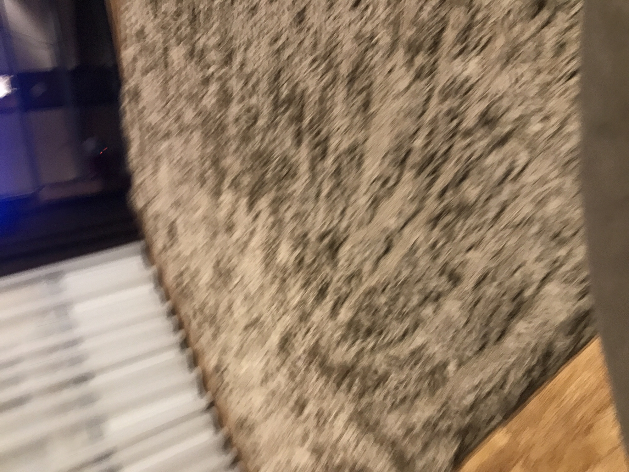 The image presents a blurry, close-up view. On the left, there is a dark object with reflective surfaces, possibly a piece of electronic equipment. Adjacent to it are parallel, light-colored lines, reminiscent of blinds or a radiator. Dominating the right side of the image is a textured, brown-grey surface, resembling a rug or carpet. A portion of a wooden surface is visible in the lower right corner. There is insufficient information to determine if this photo was taken in Yerevan, Armenia.