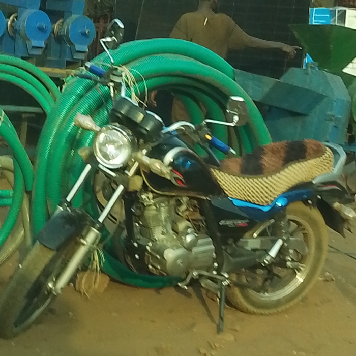 A black and blue motorcycle with a patterned seat cover stands prominently against a backdrop of coiled green hoses. To the left, blue machinery is visible. Behind the motorcycle and hoses, a man in a brown shirt stands beside a piece of blue machinery. The ground appears to be dirt.