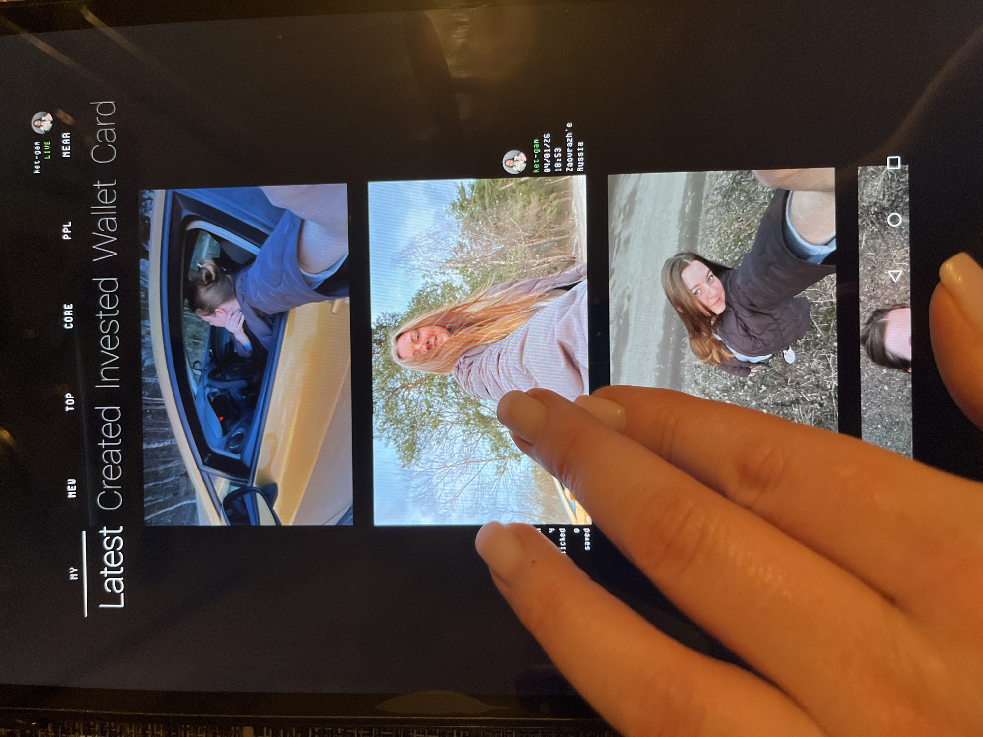 A photograph shows a hand holding a tablet. The tablet displays a social media-like feed, with multiple images of young women. One image, dated 04/01/26 18:53, indicates a location of Zaourazh'e, Russia, in Razdolye city.