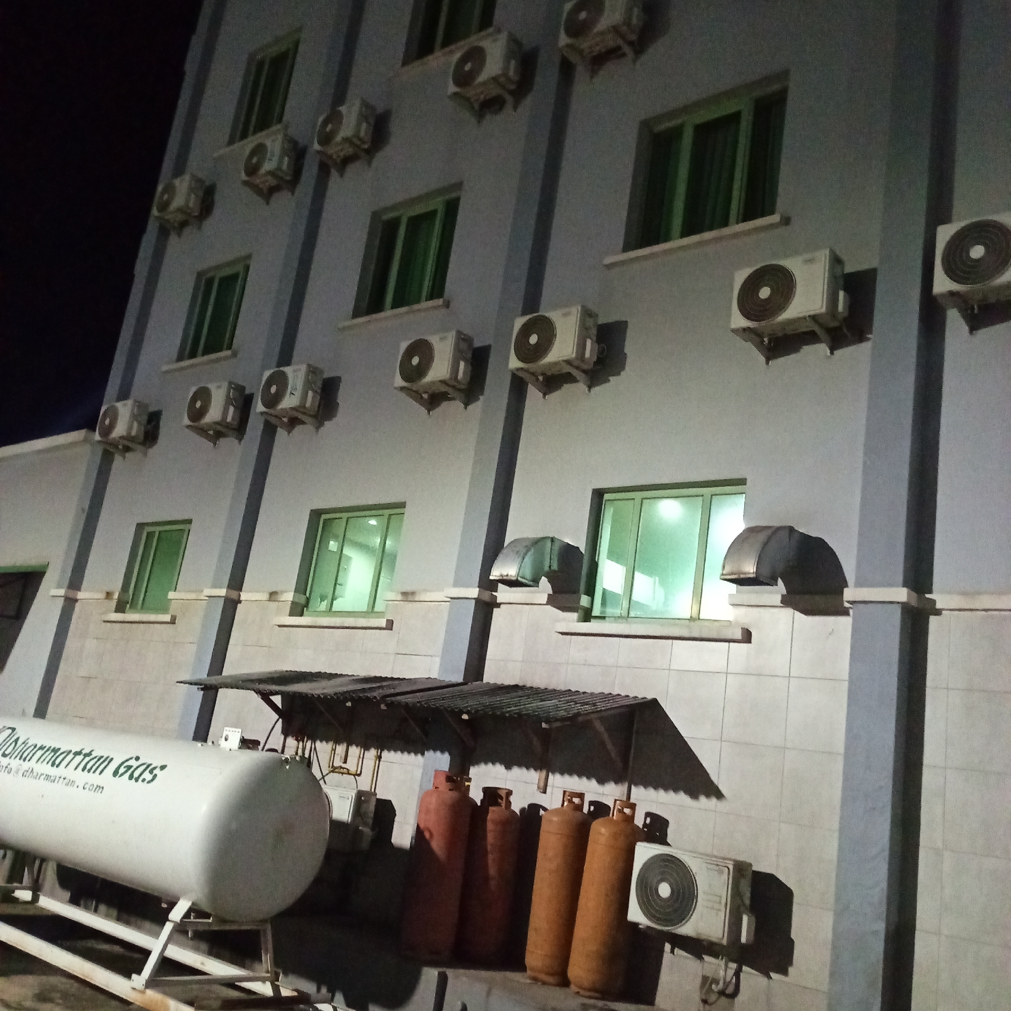 The image depicts a nighttime view of the exterior of a light blue building, likely a hotel or apartment complex, in Ibadan, Nigeria. The building has multiple floors with numerous windows, many of which are illuminated with a green glow. Mounted on the exterior walls are several air conditioning units. In the foreground, a large white cylindrical tank with the text "Dharmattan Gas" and "info@dharmattan.com" printed in green is visible, supported by a metal frame. Adjacent to the tank, several red and orange propane cylinders are arranged under a corrugated metal awning. Two more air conditioning units are visible on the lower level of the building. The scene is characterized by artificial lighting from the building's interior and the air conditioning units, with the night sky visible in the upper left portion of the frame. The overall impression is one of urban infrastructure at night, with elements related to accommodation and gas supply.
