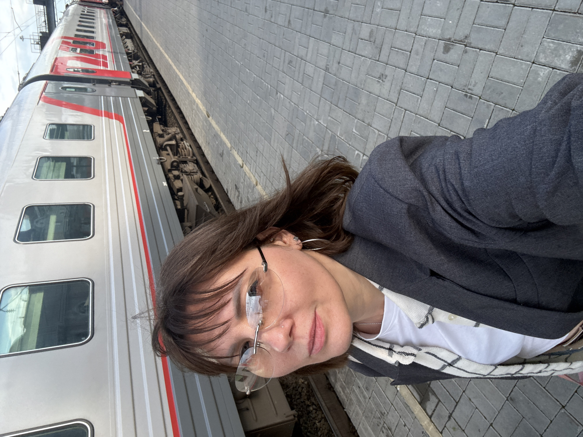 A woman with dark brown hair and bangs, wearing rimless glasses and a dark blazer over a white t-shirt and a checkered scarf, stands on a paved train platform next to a silver and red train. The train cars are visible in the background. The platform is made of grey interlocking rectangular pavers. The train has red accents on its exterior, including a diagonal stripe and markings on the doors. The scene appears to be outdoors, with the sky visible above. The image is a selfie taken by the woman, who is looking towards the camera. Based on the train markings, it is highly likely this scene is in Izhevsk, Russia, as these are typical Russian Railways train designs.