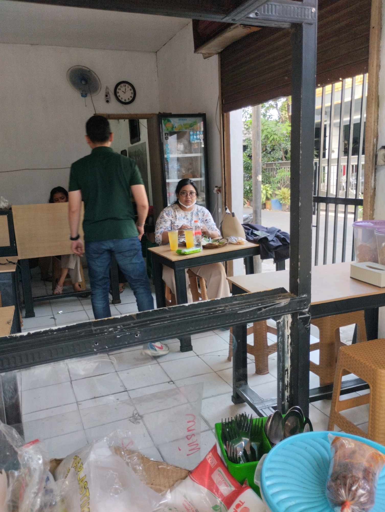 This is a forensic, matter-of-fact description of an image taken in Bekasi, Indonesia. The scene depicts the interior of a casual eatery, possibly a food stall or small restaurant, with an outdoor seating area visible through an opening.
In the foreground, the image is partially obscured by a black metal frame and a reflective surface, likely a mirror or window, showing a tiled floor with scattered plastic bags and packaging materials.
The main focus of the image is on the seating area. A man in a dark green t-shirt and blue jeans stands facing away from the camera, his back towards a woman seated at a table. Another person, partially visible, is seated behind the man. The seated woman, wearing glasses and a patterned blouse with a face mask pulled down, is looking towards the camera. She has a plate of food, a glass of orange liquid, and a bottle of water in front of her on the table.
A black and wooden table is positioned in front of the seated woman. To the left, another table is partially visible, occupied by the other individuals. Behind the seated woman, a dark jacket or fabric is draped over the back of a chair.
In the background, an electric fan is mounted on a white wall, and a clock shows approximately 2:05. A refrigerator or display cabinet with products is visible to the right of the fan. Through the opening, an outdoor environment is visible with trees, plants, and a fence with vertical bars.
The overall impression is a snapshot of a busy, informal dining establishment, possibly during daytime hours. The presence of the reflective surface in the foreground suggests the photo might have been taken through a mirror or a glass partition.