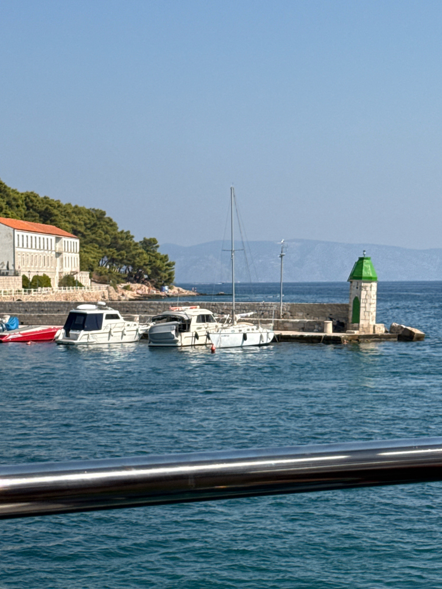A wide, vertical shot captures a harbor in Jelsa, Croatia, under a clear blue sky. In the foreground, a gleaming metal railing stretches across the bottom of the frame, obscuring the lower portion of the water. The water itself is a deep, textured blue, reflecting the sunlight.
Beyond the railing, several boats are docked. To the left, a red speedboat is tied up next to a white cabin cruiser. Another white cabin cruiser is positioned further to the right, appearing larger and more luxurious. Behind these vessels, a stone wall forms a quay.
On the quay, a small, white lighthouse with a distinctive green, conical roof stands prominently. It has a green door and appears to be made of stone. A single sailboat with a tall mast is moored to the left of the lighthouse.
In the background, a large, white building with a reddish-orange tiled roof is nestled amongst lush green trees. The building features multiple windows and architectural detailing. Further back, the landscape rises into rolling hills and distant mountains, all bathed in the bright daylight. The overall impression is one of a peaceful, sunny day in a coastal town.
