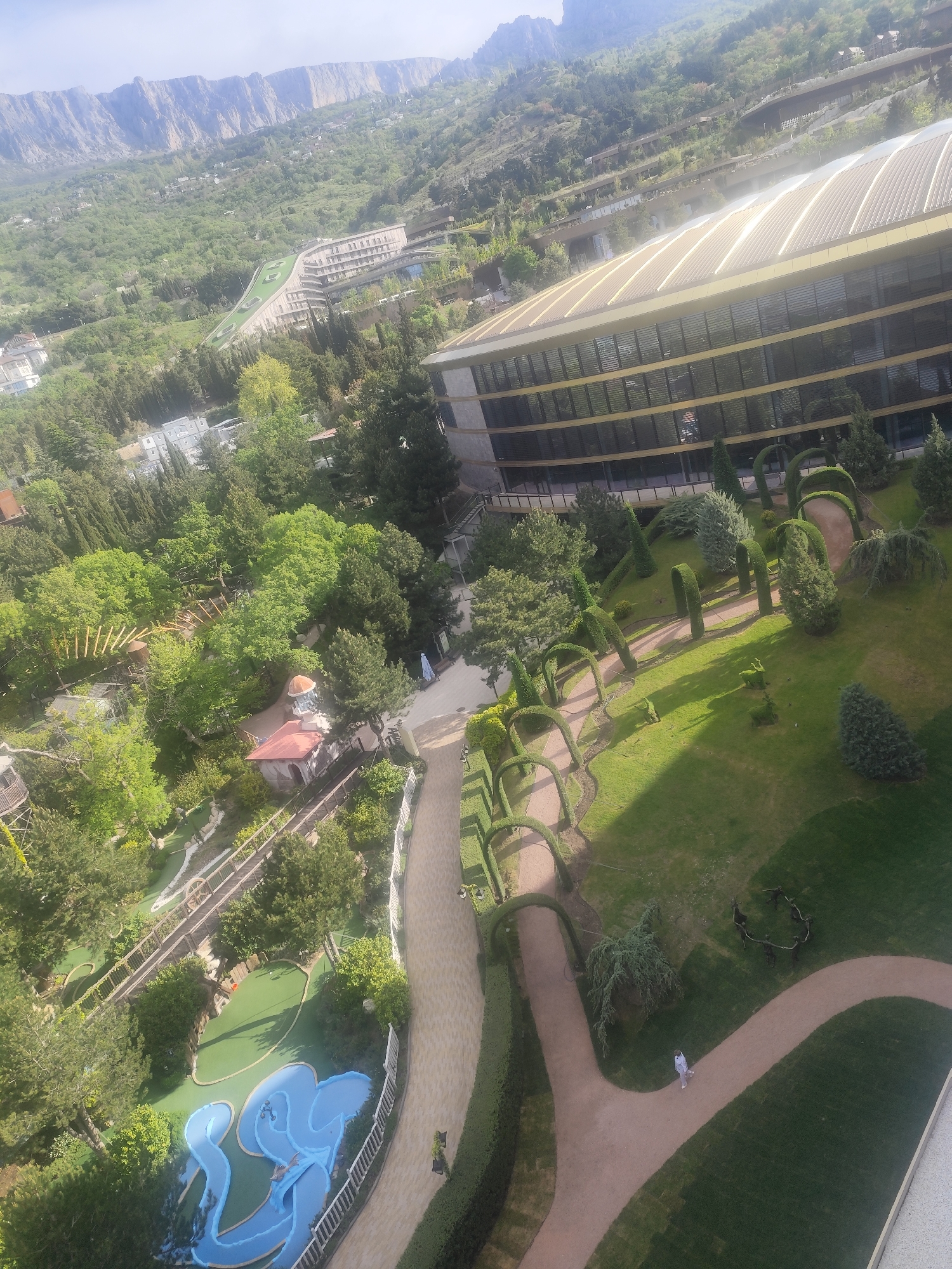 Viewed from an elevated position, the image captures a blend of manicured landscapes and natural terrains. In the upper portion, rugged, mountainous terrain meets a valley brimming with dense green vegetation. A large, modern architectural structure with a curved, reflective roofline dominates the middle-right of the frame.
Below, meticulously arranged gardens feature precisely trimmed hedges forming archways along a path. The layout creates a structured contrast to the adjacent, more untamed greenery. A miniature golf course, characterized by blue water features and green fairways, is visible in the lower left corner. Wooden structures such as bridges further add to the scene.
A solitary figure is seen walking along a winding path, suggesting human interaction within the designed landscape. In the environment, the sun's light casts long shadows, highlighting the textures and contours of the landscape. The image conveys a sense of manicured order amidst a more expansive natural setting.