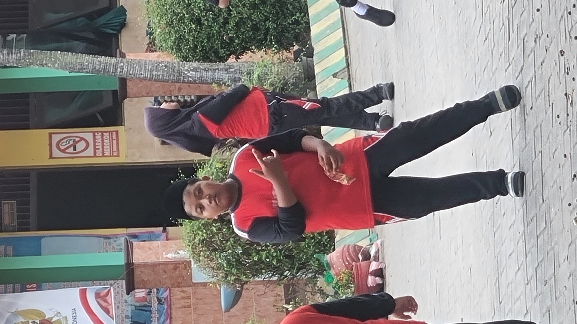 A young boy wearing a black cap, red and black long-sleeved shirt, and black pants is standing on a paved courtyard. He is holding a wrapper in his right hand and has his left hand raised with two fingers extended. He appears to be looking at the camera. In the background, a person wearing a black hijab and a red and black outfit is walking away from the camera. There is also a tree with a textured trunk and green foliage. A "No Smoking" sign is visible on a yellow wall behind them. The scene is outdoors, likely in a school or public space, given the presence of a banner with "INDONESIA" printed on it. The ground is paved with rectangular stones.