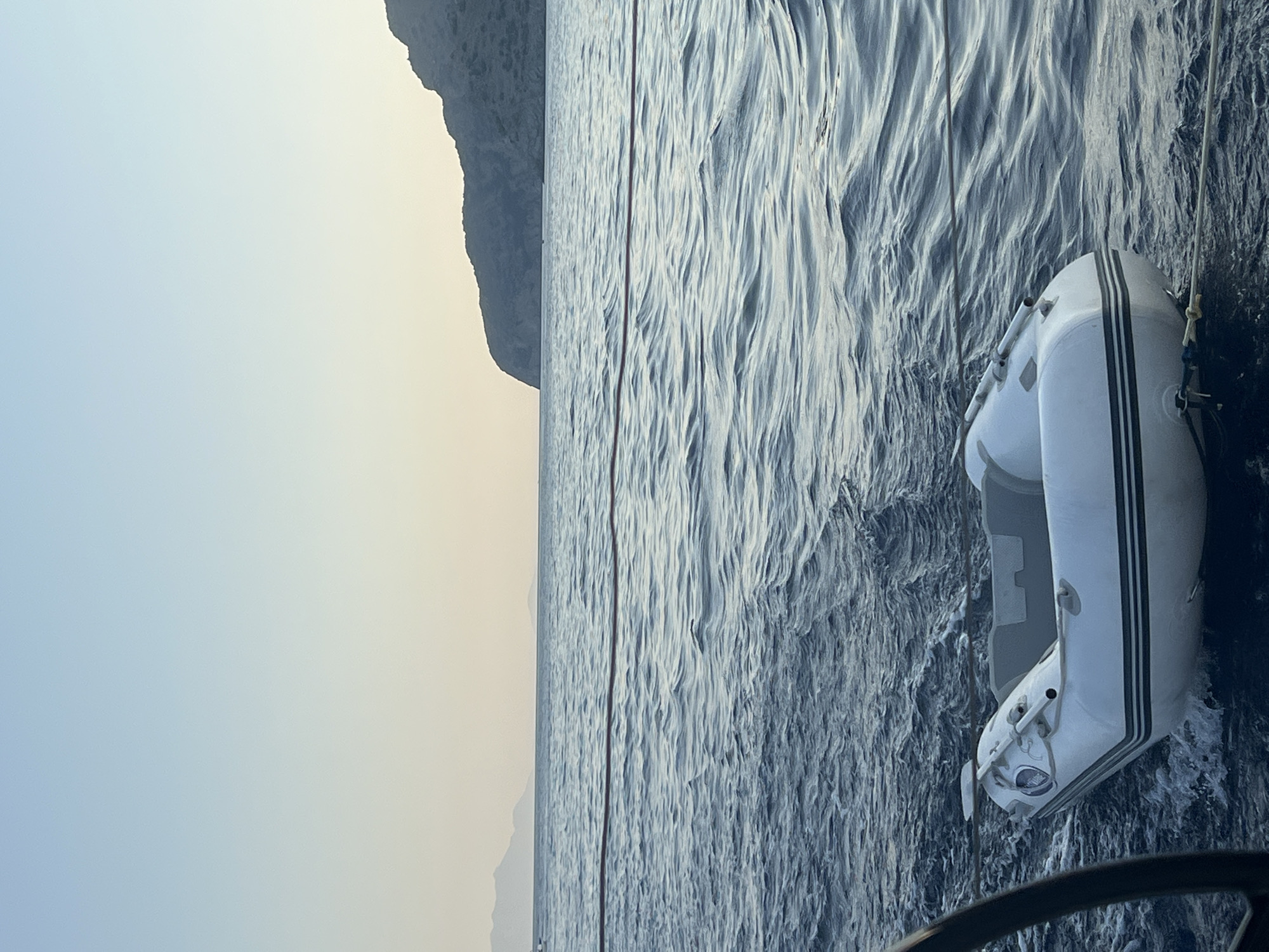 An inflatable dinghy is tethered to a larger vessel and is floating on choppy blue water under a clear, pale sky. The dinghy is white with dark grey stripes along its sides. A dark object, possibly part of the larger vessel's structure, is visible in the lower left corner. In the distance, a rocky island or coastline is visible under a hazy sky, with distant mountains on the left. The scene appears to be set at sea, possibly during sunrise or sunset due to the soft lighting and pale sky.