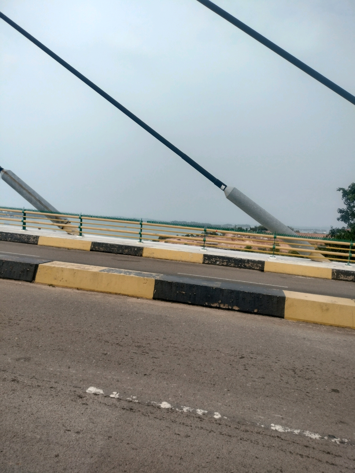 The image depicts a section of a cable-stayed bridge in Batam, Indonesia, likely over a body of water. In the foreground, there is an asphalt road with black and yellow concrete barriers. The road shows some wear and tear, with white markings and scattered debris.
The bridge structure is visible with large, gray cylindrical supports angling upwards, connected to dark cables that extend towards the top of the frame. These cables are a prominent feature, creating diagonal lines against the overcast sky.
Behind the road and barriers, there is a yellow and green railing system along the edge of the bridge, separating the roadway from a lower section. This lower section has a lighter-colored surface and appears to be part of the bridge deck or approach. Further in the distance, a hazy landscape is visible, suggesting water and possibly the coastline of Batam. A few trees are on the right side of the image, providing a touch of greenery. The overall impression is one of an infrastructure project in a coastal or riverside environment under a cloudy sky.