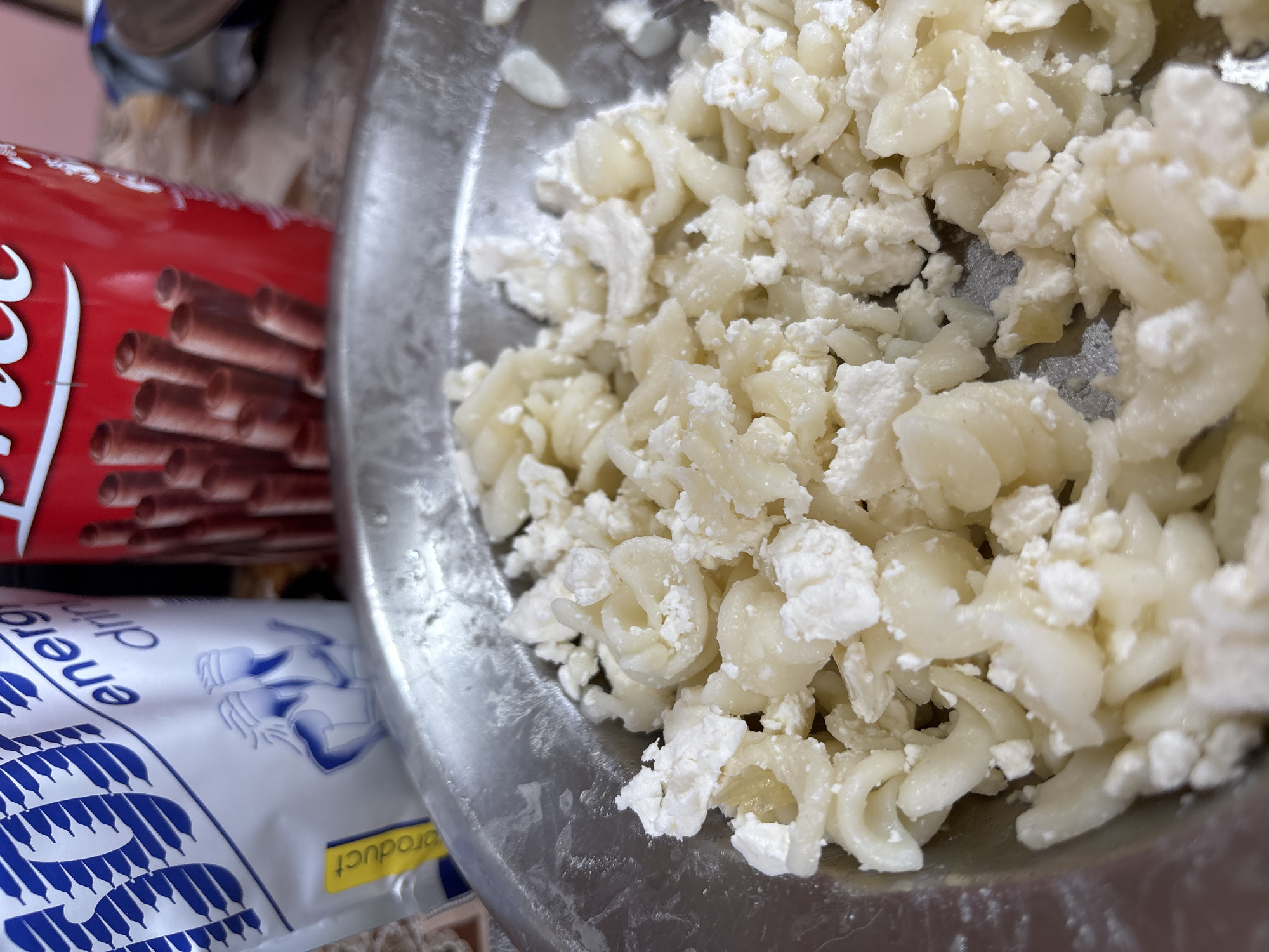 This close-up image, taken in Krivodol, Bulgaria, features a metal bowl filled with a mixture of food, flanked by two different packaged items.
The main subject is the food in the shiny metal bowl, which consists of small, pale yellow, curved pasta shapes (resembling macaroni) mixed thoroughly with white, crumbly cheese, similar to cottage cheese or feta. The texture suggests a soft, prepared dish, possibly macaroni with cheese, that is ready to be consumed or further prepared.
To the left of the bowl, a vibrant red cylindrical packaging is partially visible. It displays the characteristic "Coca" script, indicating the Coca-Cola brand, and features an image of several brown chocolate-filled wafer sticks or biscuit rolls.
Below the red packaging, a white and blue plastic bag or wrapper is also partially seen. Visible text includes "energ dran," likely an abbreviation for "energy drink," and "product" on a small yellow label. The bag features a pattern of interconnected blue shapes, possibly an abstract representation of energy or a stylized logo.
The scene appears to be an indoor setting, likely a kitchen or dining area, with casual lighting. There is no specific activity depicted other than the arrangement of these items, suggesting a paused moment before a meal or snack. The overall impression is one of a simple, domestic food preparation or mealtime setting.