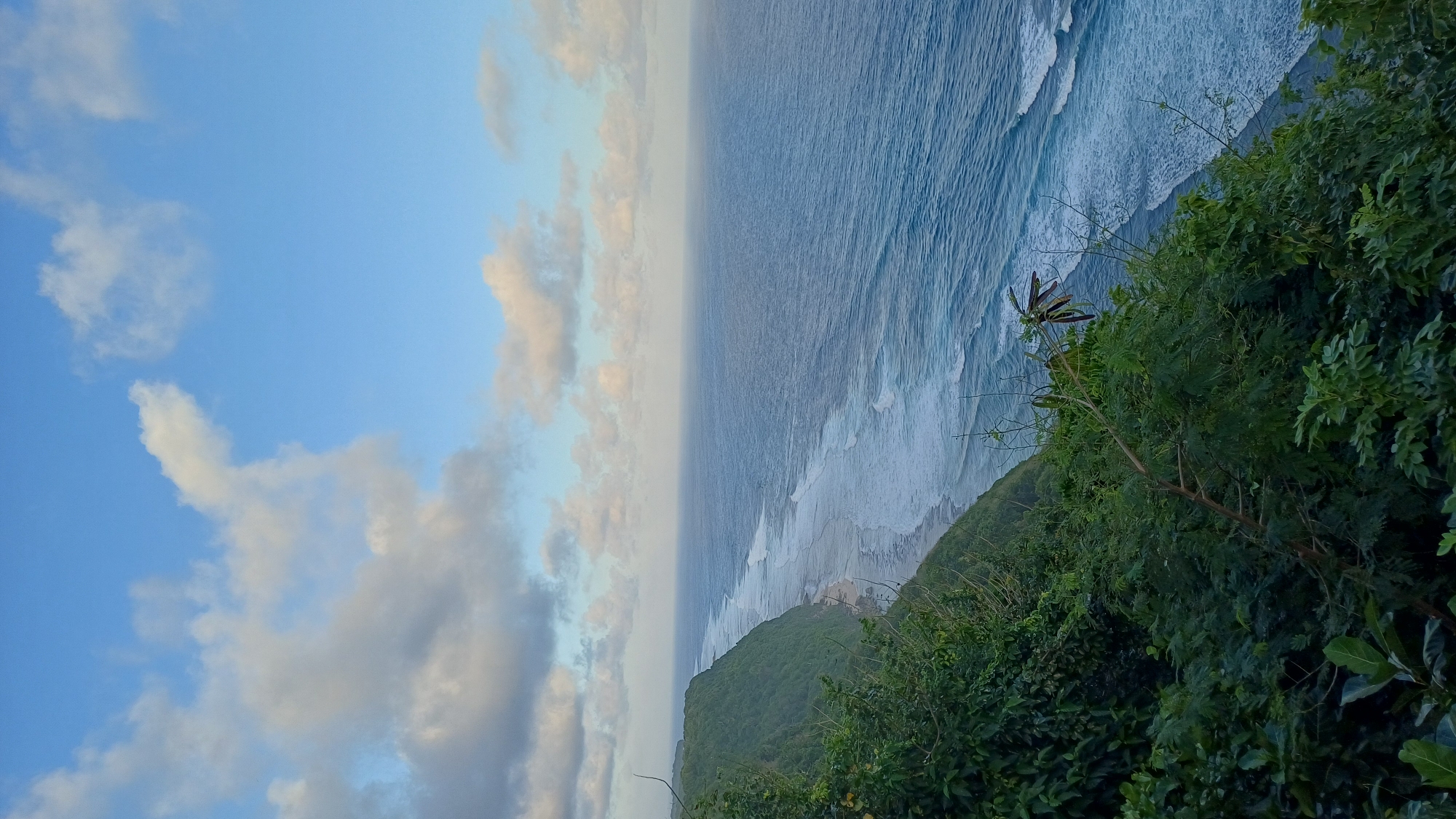 The image shows a coastline in Kuta Selatan, Indonesia. The scene features a view from a cliff overlooking the ocean. The sky is partly cloudy with hues of blue and soft white clouds. The ocean is a gradient of deep to light blue, with visible waves approaching the shore. A line of lush green vegetation covers the cliffside, extending down toward the beach. There are no people visible in the image.