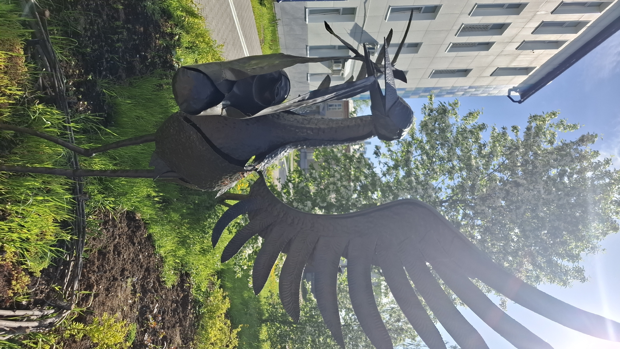The image shows a metal sculpture of a mythological bird with wings, standing on a grassy area near a sidewalk. The sculpture has a long neck, a stylized head with several horn-like projections, and large wings. In the background, a multi-story building with multiple rows of windows can be seen, along with a tree in full bloom. The location is believed to be in Chaikovsky, Russia. The sky is blue and the sun is shining.
