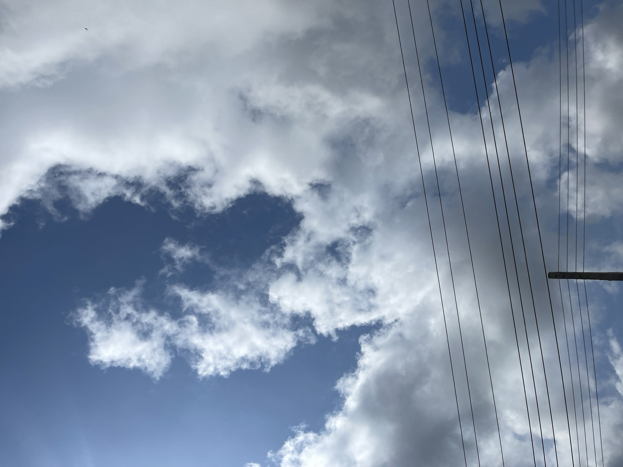 This image captures a clear blue sky partially obscured by scattered white clouds. Several parallel power lines run horizontally across the lower half of the frame, with a single wooden utility pole visible in the bottom right corner. A faint dark speck, possibly a bird or insect, is visible in the upper right portion of the sky. The scene appears to be an outdoor view, possibly from a residential or commercial area, given the presence of power lines and a utility pole. The lighting suggests it is daytime, with sunlight filtering through the clouds. The general atmosphere is one of an ordinary day, likely in a place with overhead electrical infrastructure. The image does not provide any specific visual cues to pinpoint its location within Jos, Nigeria, beyond the context provided.