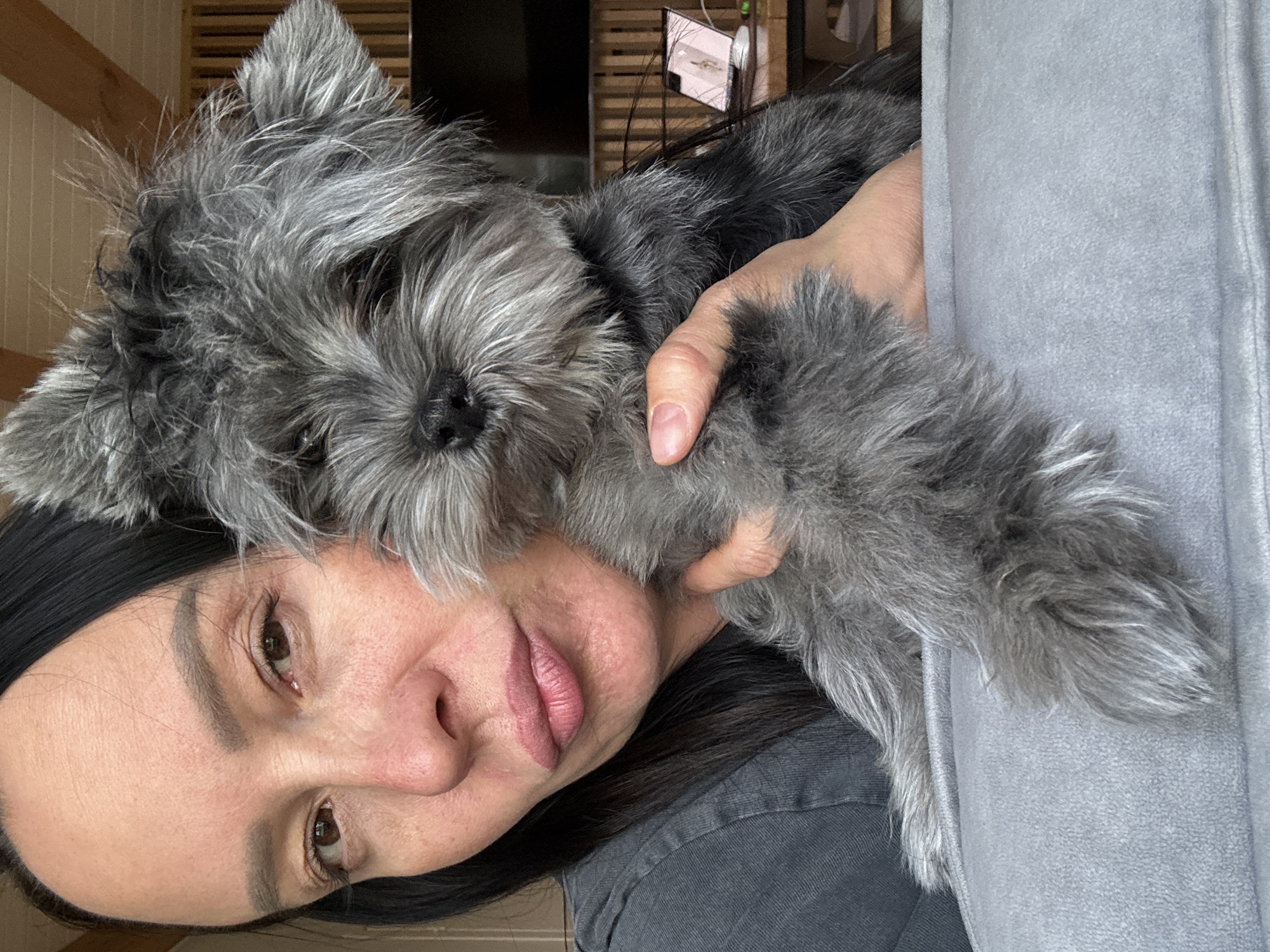 A woman holds a gray, furry dog close to her face. The dog is positioned on her shoulder and chest area. The woman wears a dark-colored shirt and a headband in her dark hair. The background includes a wooden-slatted wall and a glimpse of a television. The location is Chaikovsky, Russia.