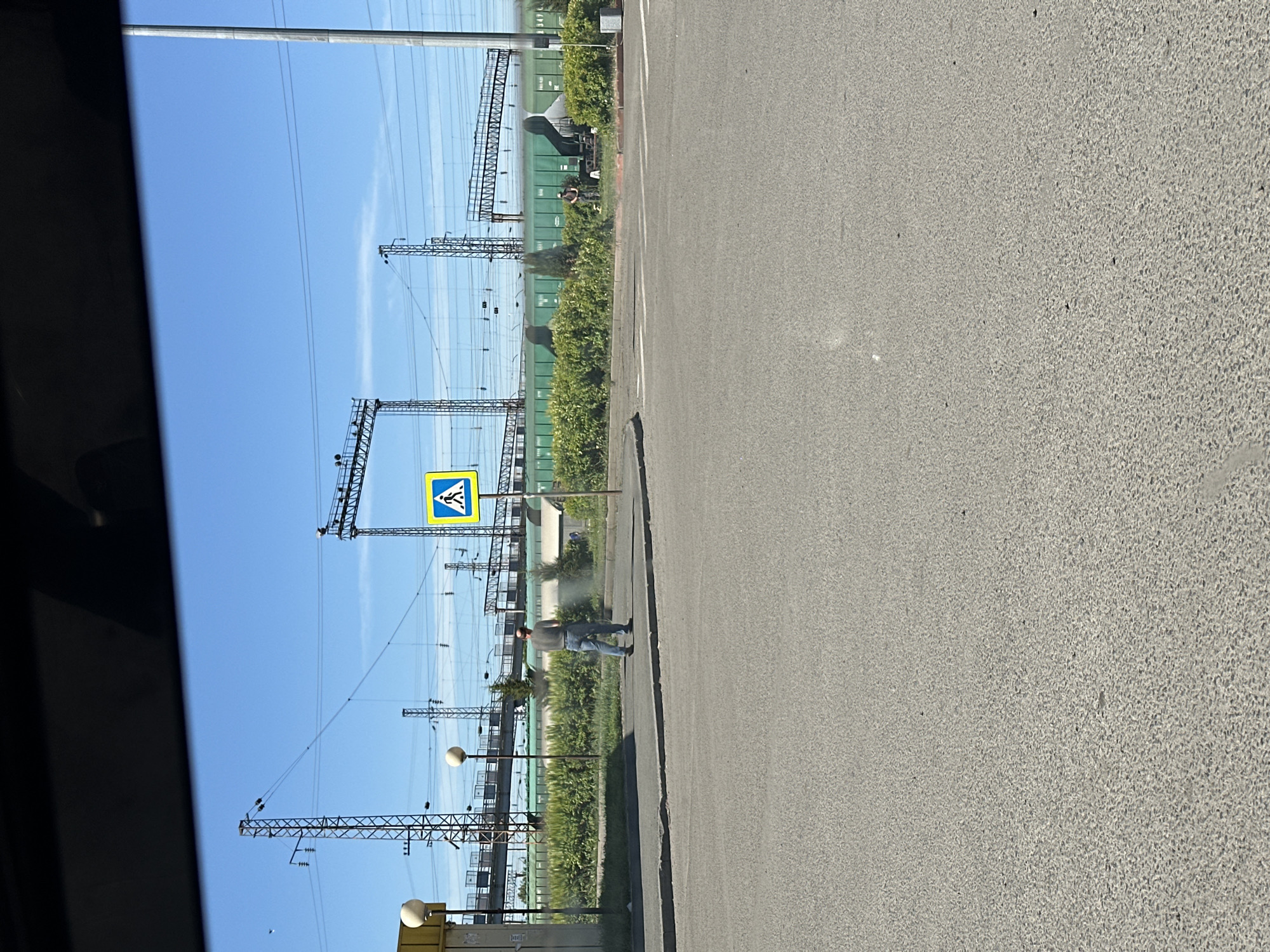 The image shows an outdoor scene at what appears to be a pedestrian crossing near a railway line in Novoaltaysk, Russia. In the foreground, there is a paved road with white markings indicating a crossing. A man in a dark shirt and jeans is standing on the sidewalk, facing away from the camera, possibly watering some plants with a hose that is spraying a mist. Behind him, there is a line of green train cars on a railway track, with overhead power lines and support structures visible. A yellow pedestrian crossing sign with a black silhouette of a person walking is placed on a pole to the right of the man. The sky is clear and blue. In the background, there are several large industrial structures and green vegetation. Further down the track, two people are sitting near the train cars. The overall environment suggests an industrial or transportation hub area.