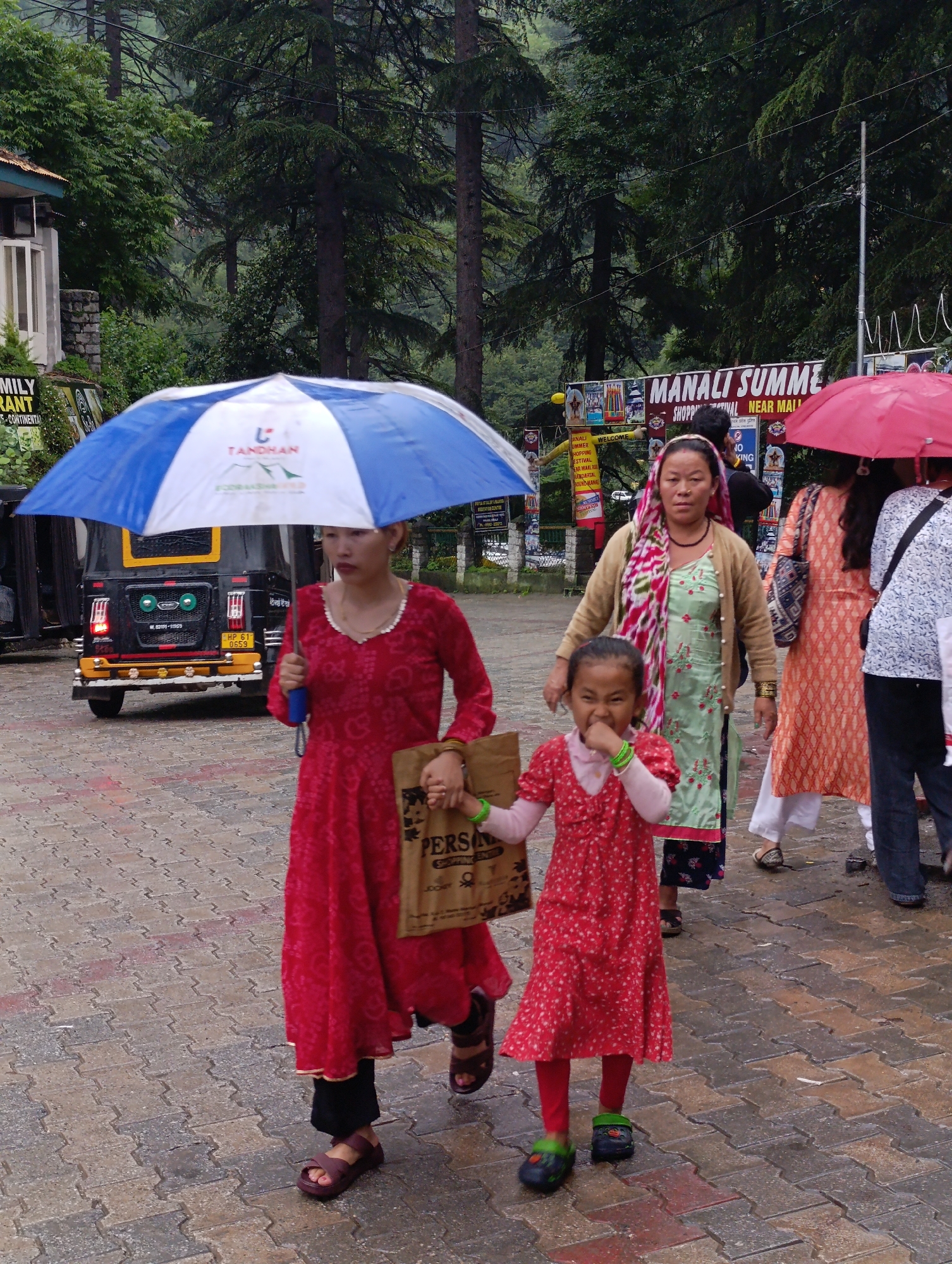 In Manali, India, on a paved street under a cloudy sky, a group of people walk together. A woman in a red dress walks under a blue and white umbrella, carrying a brown paper bag. Beside her, a young girl in a red dress with white polka dots walks, holding the woman's hand and looking at the camera with her hand near her mouth. Behind them, another woman in a green and pink outfit and a beige cardigan walks. To the right, a woman in an orange and white patterned dress, and a woman in a white and blue patterned shirt walk. A red umbrella is visible in the background. An auto-rickshaw is parked on the left side of the street. Signs in Hindi are visible in the background, including one for "MANALI SUMMER SHOPPING FESTIVAL". The environment is lush with trees in the background.
