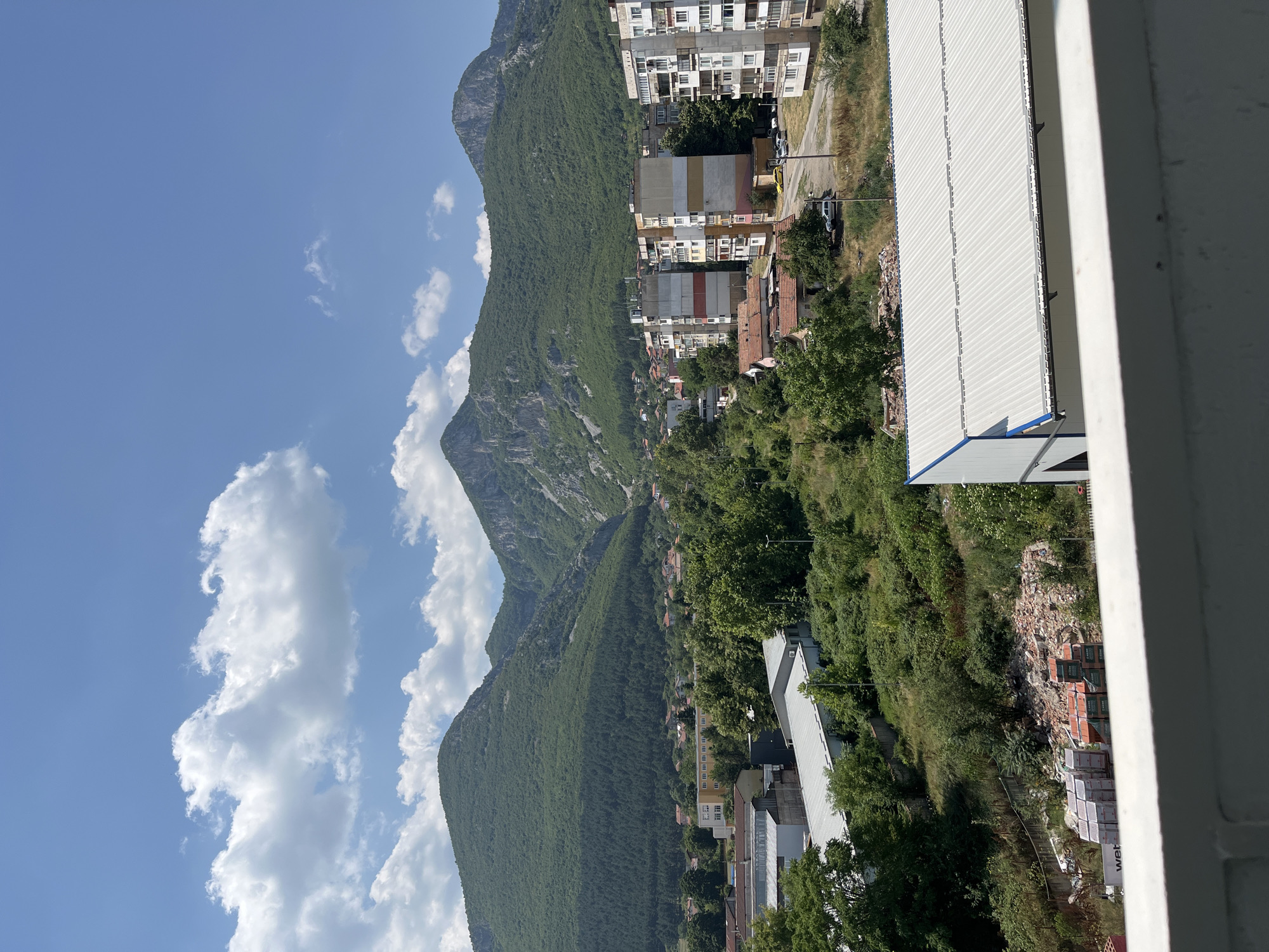 The image displays a panoramic view of a town nestled at the foot of a mountain range under a clear blue sky with scattered white clouds. The foreground is dominated by a white railing and the roof of a modern building. The middle ground reveals a mix of urban and natural elements. Several multi-story apartment buildings with varying architectural styles and colors are visible, some appearing older with balconies and others more modern. Scattered among the buildings are smaller residential houses with red-tiled roofs. Green trees and vegetation are abundant throughout the town, suggesting a lush environment. Industrial or commercial buildings with metal roofs are also present. A road winds through the town, with a few cars parked or driving. In the background, the imposing green mountains rise, densely covered with trees, with rocky outcrops visible in some areas. The overall impression is of a town that harmoniously blends with its natural mountainous surroundings. This scene is characteristic of the city of Vratsa, Bulgaria, known for its picturesque location against the backdrop of the Vratsa Mountains.