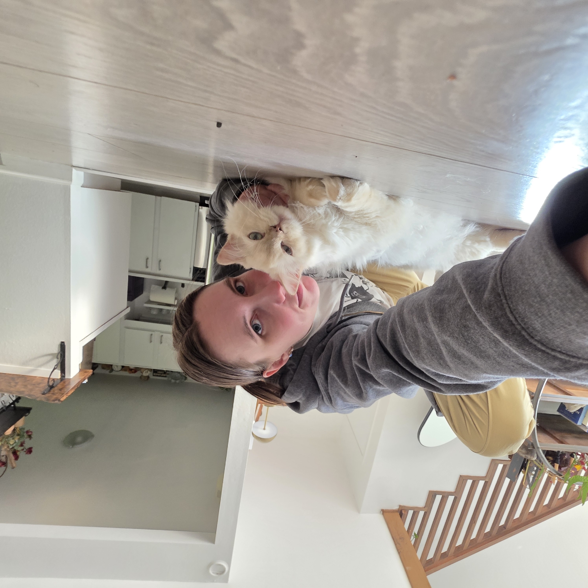 In Portland, Oregon, a person with short brown hair, wearing a grey hooded sweatshirt and yellow pants, is lying on a light-colored wooden floor. They are taking a selfie with a fluffy white cat. The person is smiling and looking at the camera, while the cat is lying down next to them, also looking at the camera. The background shows a kitchen with white cabinets and countertops, and a wooden staircase with a railing. The scene appears to be indoors in a residential setting.