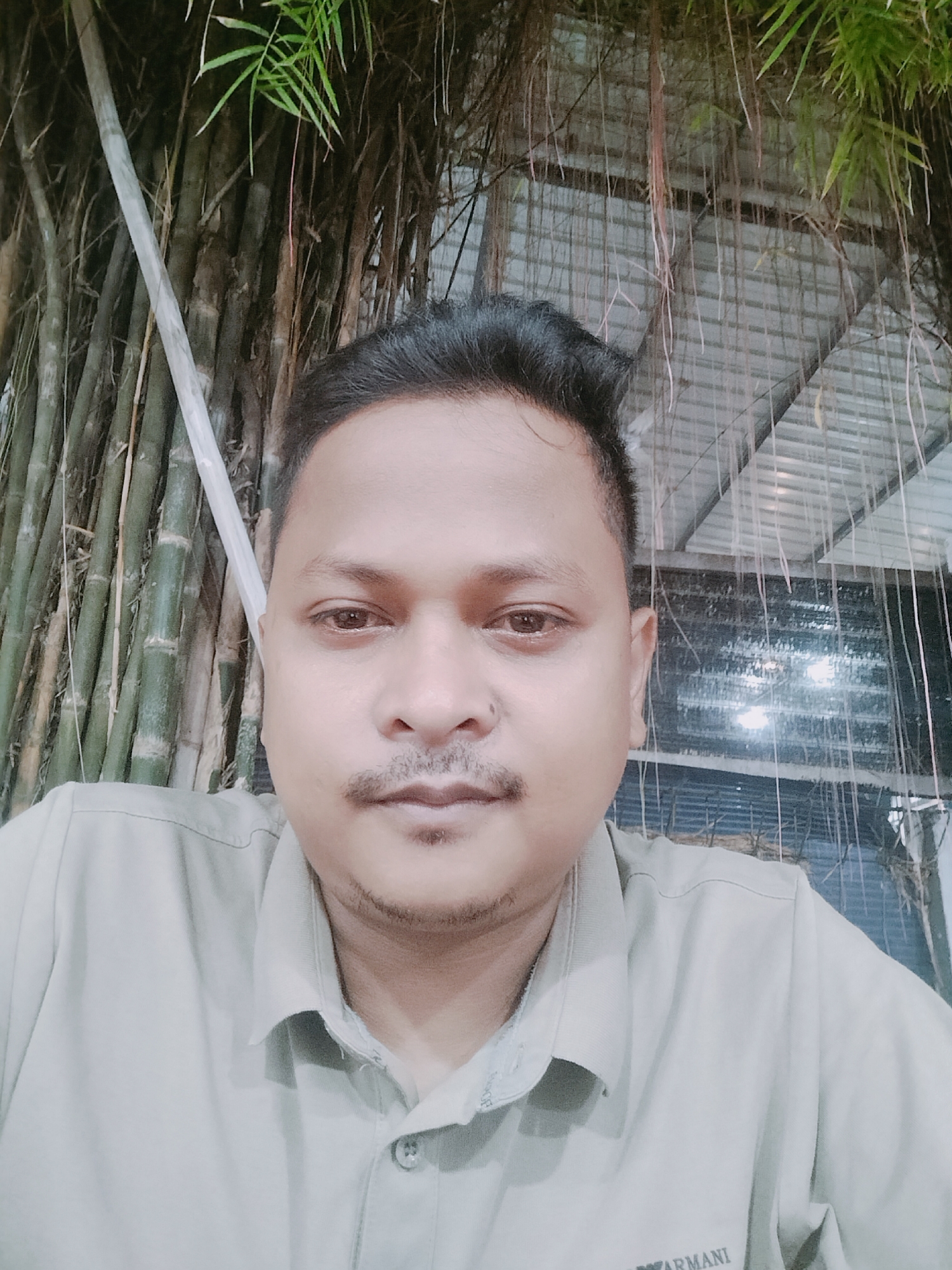 The image is a close-up selfie of a man in Lhokseumawe, Indonesia. The man has dark hair and a mustache. He is wearing a light-colored collared shirt. In the background, there is a cluster of bamboo stalks and hanging vines. Above the man's head, a corrugated metal roof is visible, with some lights reflecting on its surface. The overall lighting is dim, suggesting it might be evening or indoors.