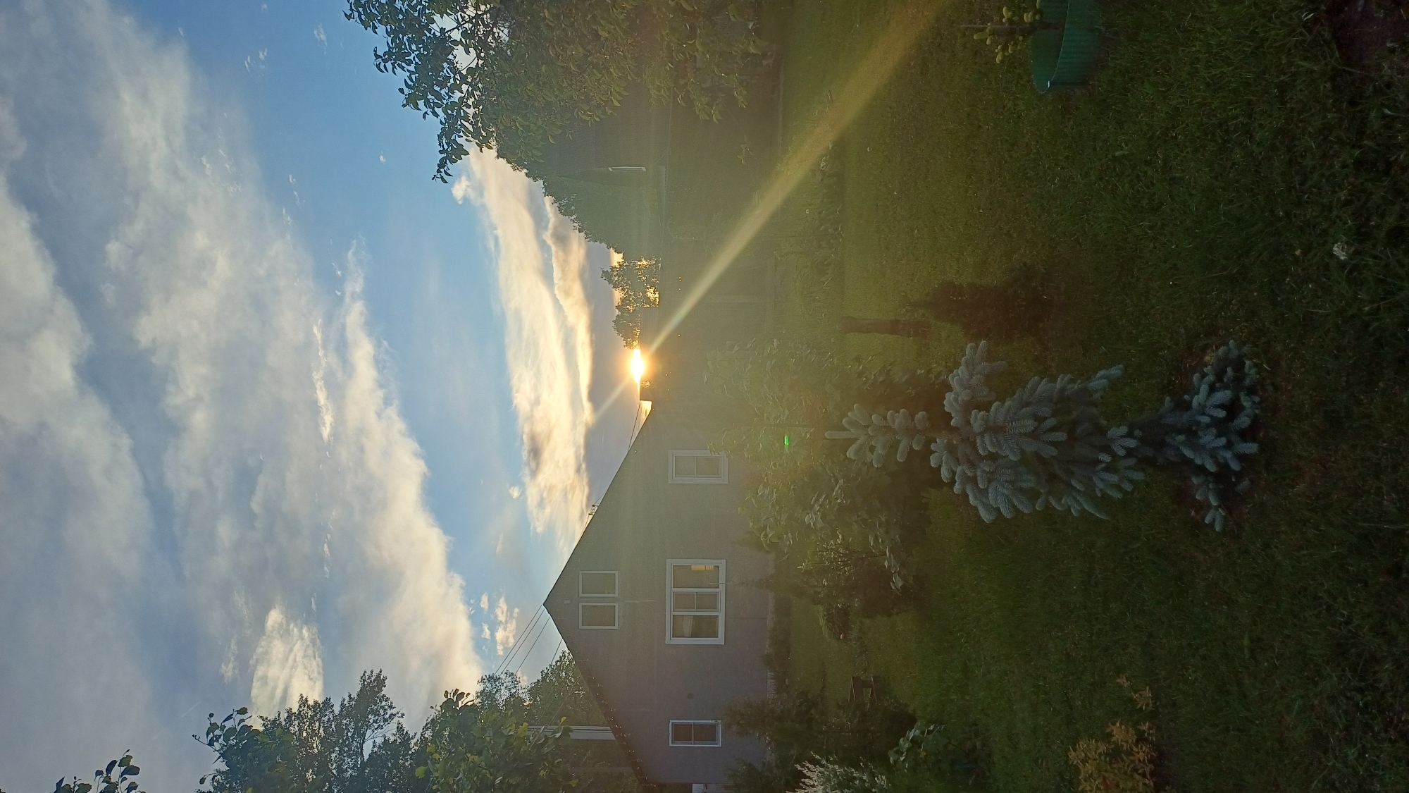 A low-angle, wide shot captures a landscape dominated by a house, sky, and vegetation, likely in a rural area. The house, grey with white-framed windows, occupies the lower-center of the frame. The sun, positioned directly above the peak of the roof, casts a long, bright beam downward. To the left, the sky is blue, partially covered by streaks of white clouds. Trees are positioned along the top and left of the frame. To the right of the house, the ground is covered in green grass, with a blue-green shrub visible. The lighting suggests either sunrise or sunset. There are no visible people or interactions. No specific information confirms the location is Timokhovo, Russia.