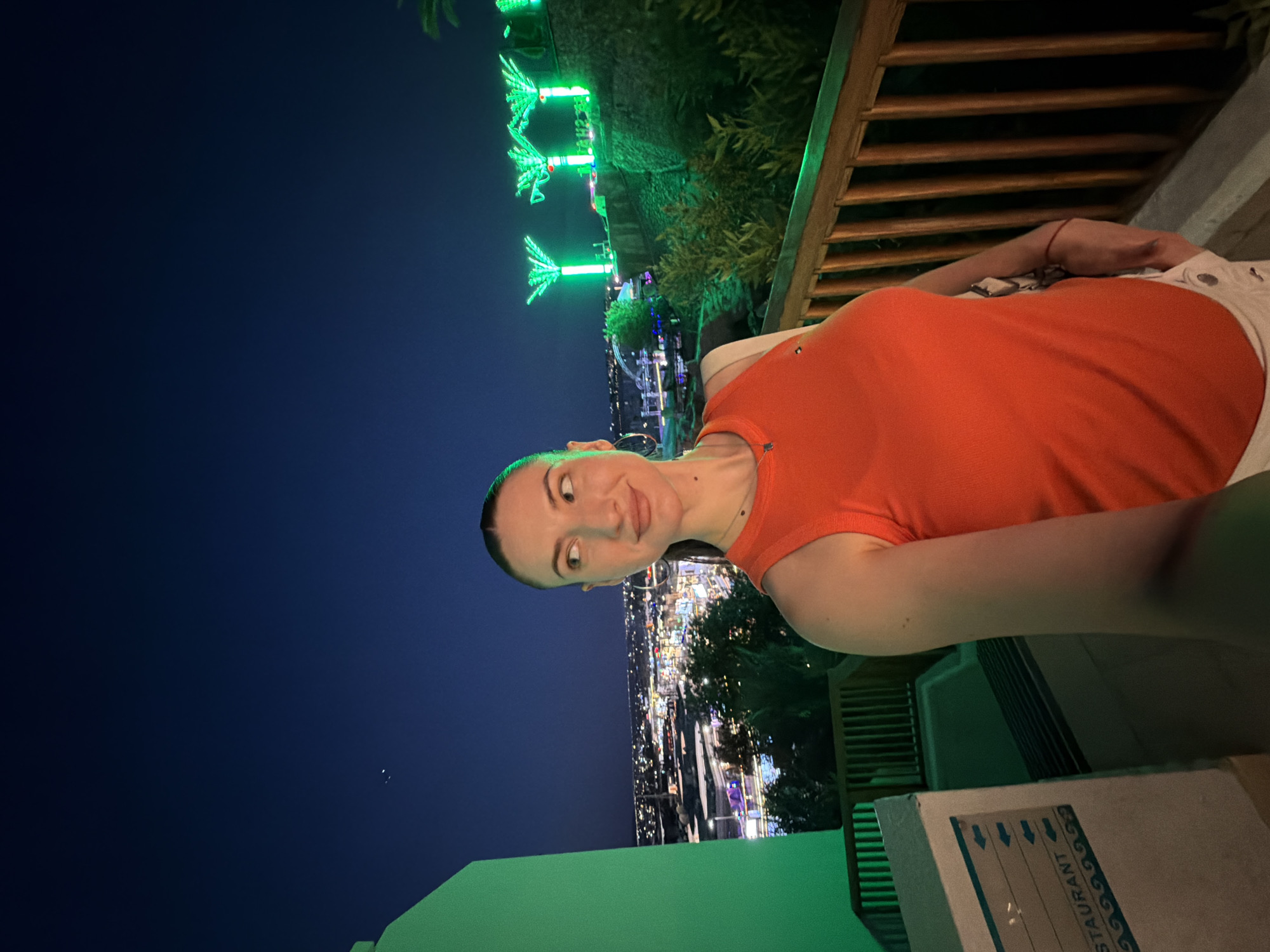 A young Caucasian woman with dark hair pulled back stands on a balcony at night, looking to her left. She is wearing an orange sleeveless top and white shorts. The background features a city skyline illuminated by numerous lights, suggesting an urban setting. To the right, stylized green palm tree-shaped lights are prominent. The woman's pose and the framing of the shot suggest she is taking a selfie. The environment appears to be a resort or hotel overlooking the city of Sharm El Sheikh, Egypt, given the distinctive lighting and palm tree motifs often associated with tourist areas in that region.