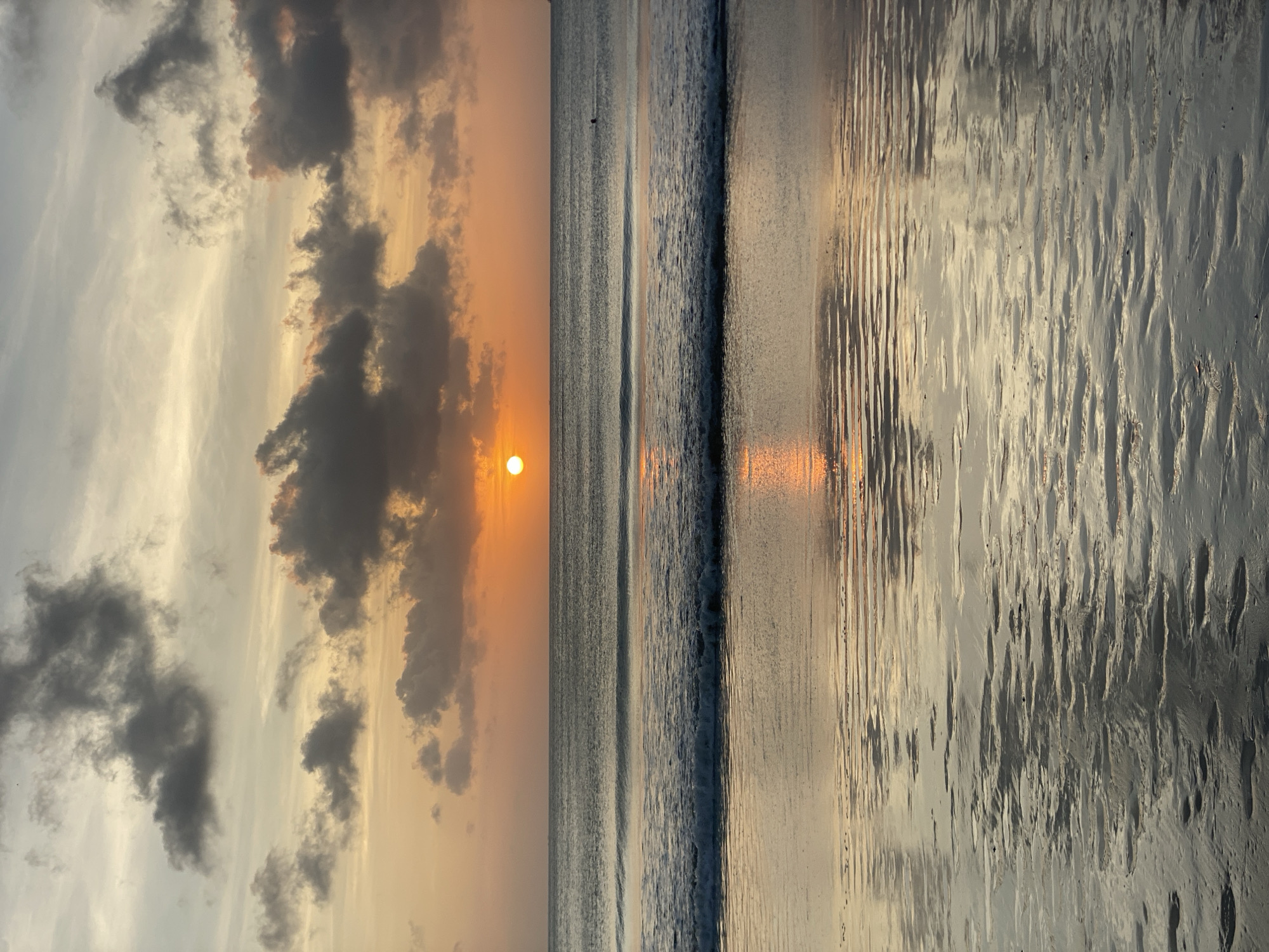 The image captures a sunset scene at a beach in Kuta Selatan, Indonesia. The sun is visible, partially obscured by clouds, casting an orange glow across the sky and reflecting on the wet sand and ocean surface. There are gentle waves along the shoreline. The beach appears relatively deserted, with no immediately discernible people or subjects clearly visible in the frame. The sand shows patterns, likely due to receding water. The overall scene conveys a serene and calm atmosphere.