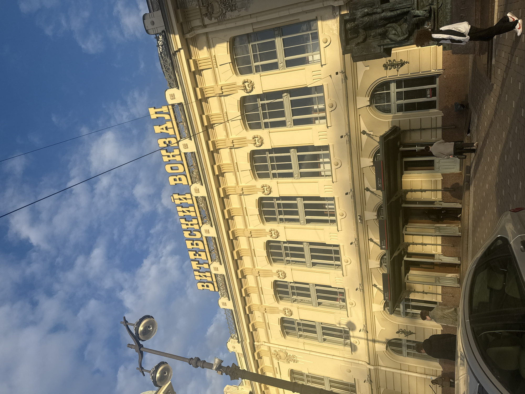 The image provides a tilted, low-angle view of the ornate facade of the Vitebsky Railway Station in Saint Petersburg, Russia, under a partly cloudy blue sky. The grand building is a pale yellow or cream color, illuminated by natural daylight which casts soft shadows, suggesting it is not midday.
Architecturally, the station features multiple stories with large, gridded rectangular windows on the upper floors and arched windows or entrances on the ground level. The facade is richly decorated with classical elements, including pilasters, cornices, and sculptural details such as lion-head carvings between the window levels. High on the building, a prominent sign in bright yellow Cyrillic letters spells "ВИТЕБСКИЙ ВОКЗАЛ," which translates to "Vitebsky Railway Station."
Several individuals are visible on the paved sidewalk in front of the building. One person with dark, curly hair, wearing a white and black jacket, black pants, and white and red sneakers, stands beside a large, dark, multi-figure sculpture. Another person, seen from the back in a light jacket, walks towards one of the station's arched entrances. Further down, a person in a dark jacket and other indistinct figures can be observed. Part of a light-colored car is visible on the street to the right. In the foreground on the left, a traditional street lamp with multiple ornate light fixtures stands against the sky. The scene captures everyday activity around this historic urban landmark.