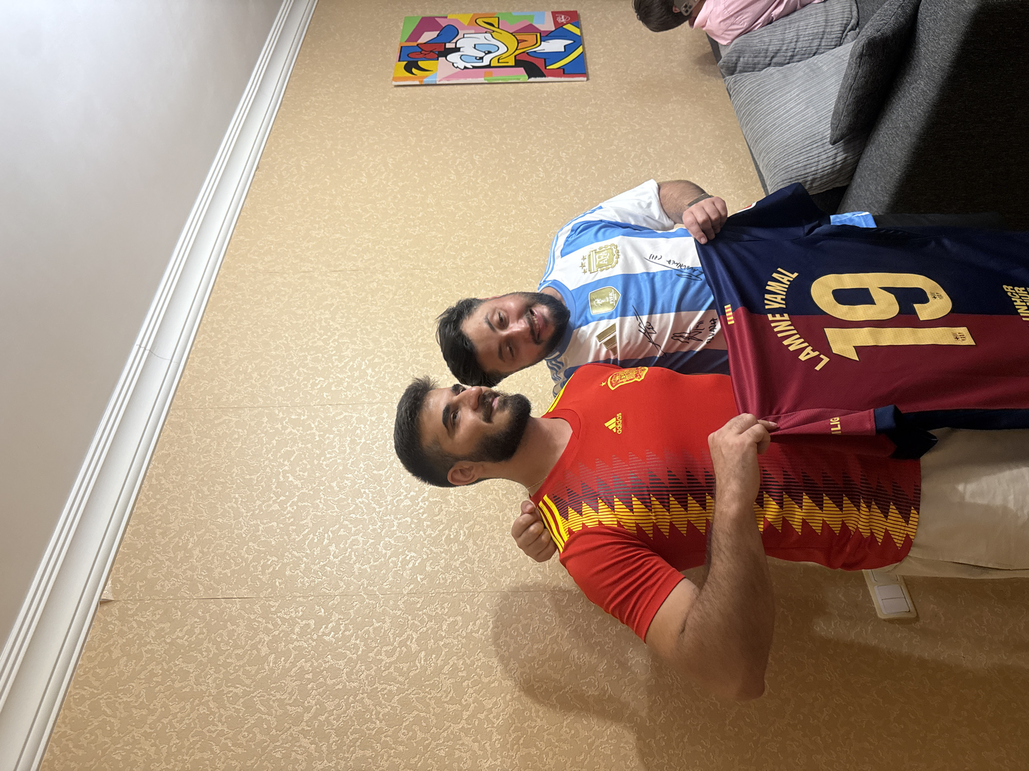 Two men are posing for a photo indoors. The man on the left is wearing a red Spain national football team jersey. The man on the right is wearing a light blue and white striped Argentina national football team jersey. Both men are smiling and holding up a burgundy and navy blue football jersey with the name "LAMINE YAMAL" and the number "19" printed on it in gold lettering. The background features a textured, light brown wallpaper and a framed picture of Donald Duck on the wall. A sofa is visible on the right side of the image. The scene appears to be in a residential setting. There is no specific indication of the location in the city of Yerevan, Armenia.