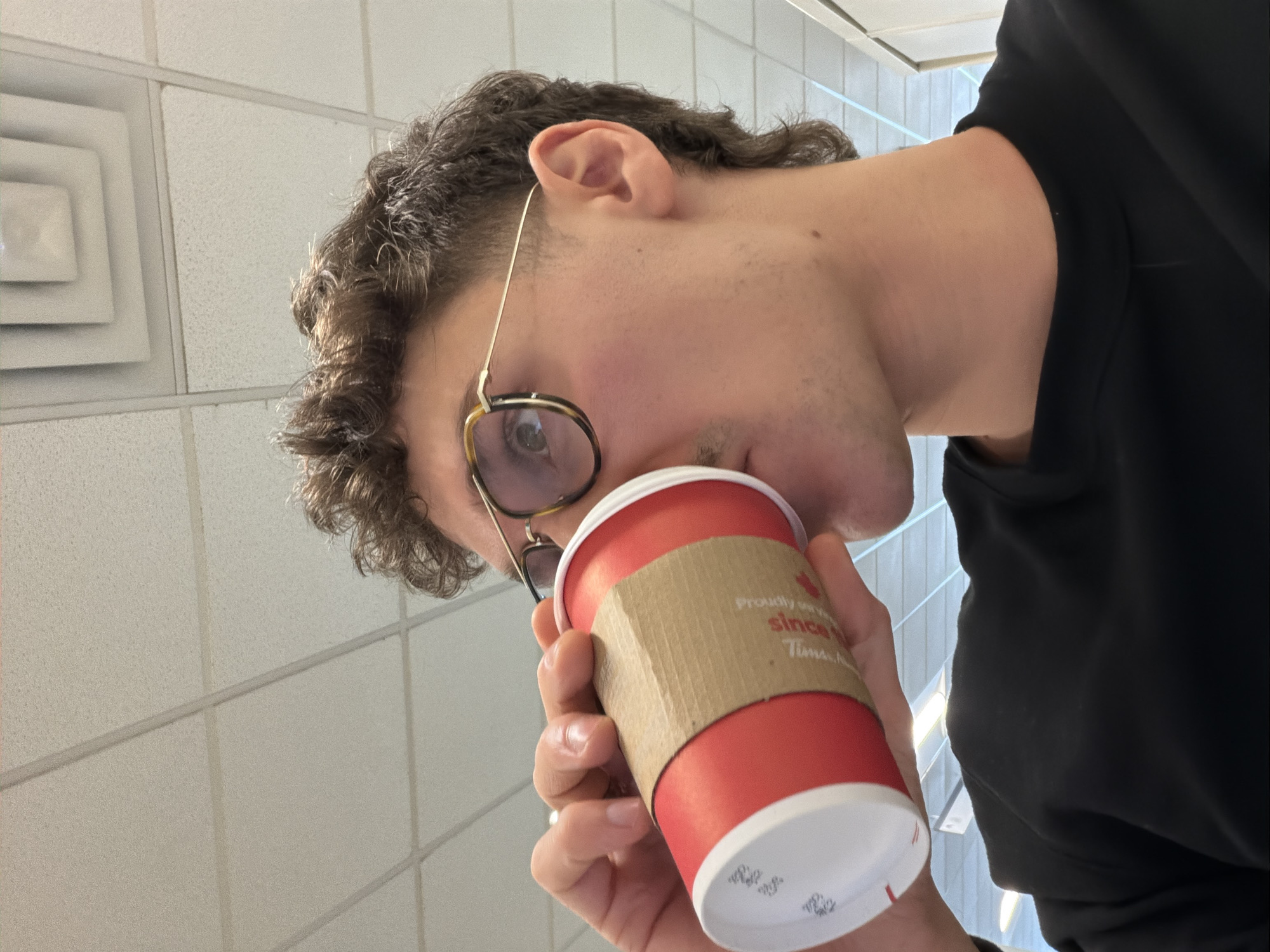 A male individual is depicted in a close-up, profile view, drinking from a red Tim Hortons disposable cup within an indoor commercial or institutional setting in Mirabel, Canada. The subject has short, curly brown hair styled in a short mullet, a light mustache, and wears gold-framed eyeglasses with tortoiseshell accents. His right hand grips the cup, which features a beige corrugated sleeve and a white lid. The background consists of a white drop ceiling with visible grid lines, acoustical panels, and a multi-layered square air vent.