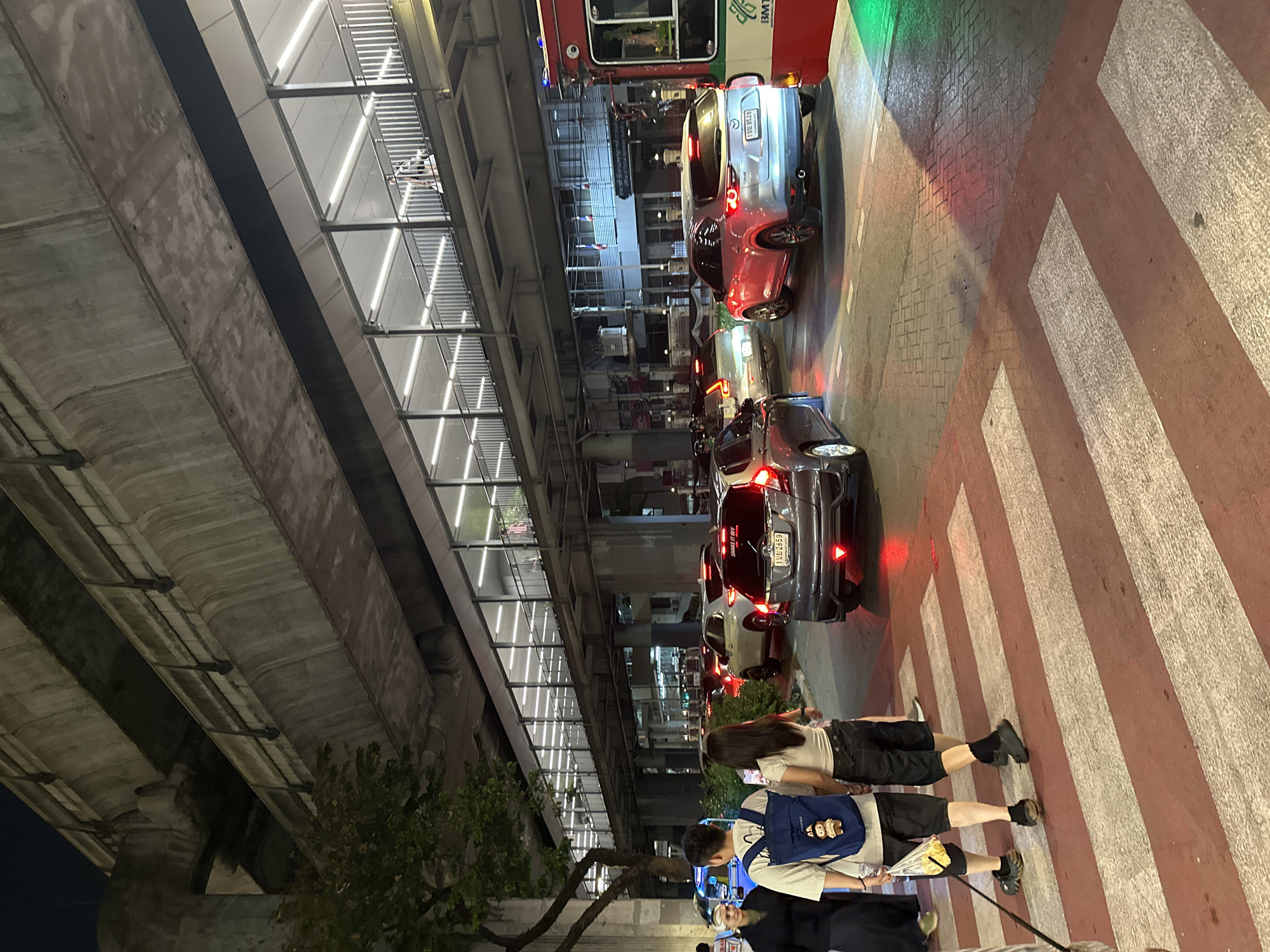 The image shows a street scene in Bangkok, Thailand, at night. An elevated train track runs along the left side of the image, with a pedestrian walkway above the street. Several vehicles are lined up in traffic, including cars and a bus, on the right side of the image. People are walking across a pedestrian crosswalk in the foreground. The environment is illuminated by streetlights and vehicle lights.