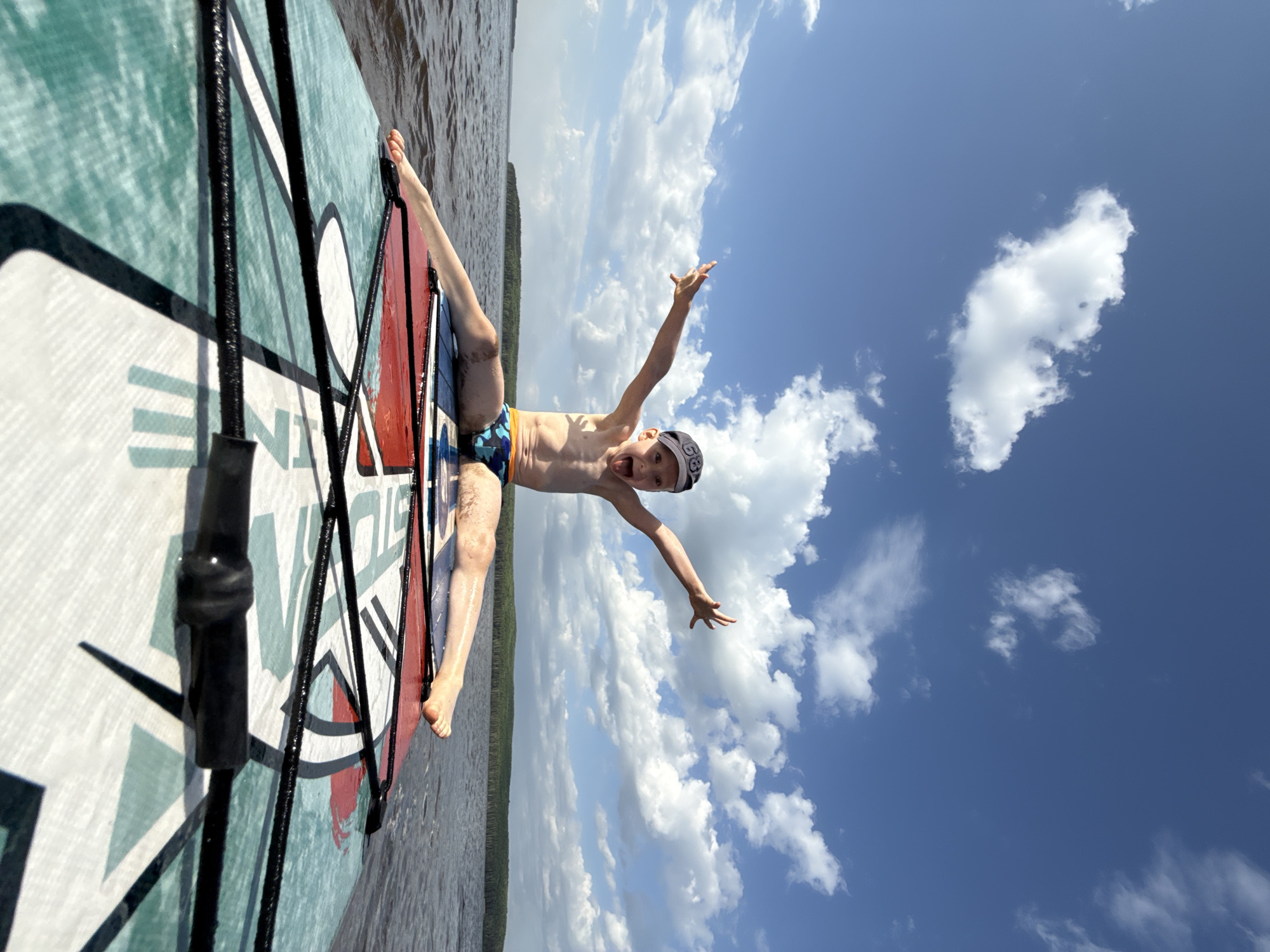 A boy is standing on a windsurfing board on a body of water with a treeline in the background and a bright blue sky with scattered clouds. The boy is wearing a blue and orange swimsuit and a backwards baseball cap. He is standing with his legs spread and arms outstretched, yelling and smiling at the camera. The windsurfing board is teal, white, and red. The water is dark and reflects the treeline. The treeline appears to be far off in the distance. It is daytime. The photo was likely taken in Chaikovsky, Russia.