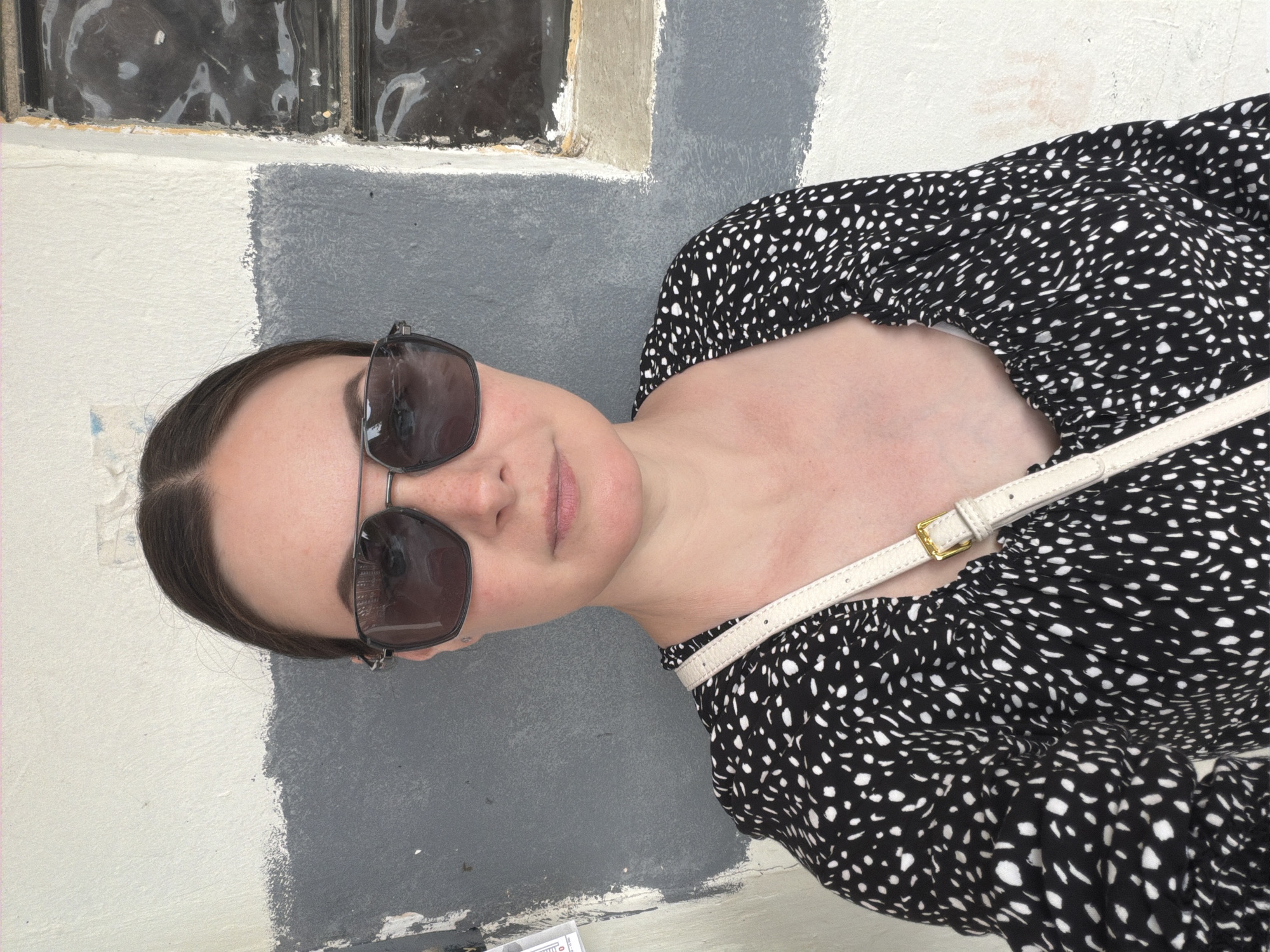 A woman with sunglasses is standing against a painted wall. The wall has an old window near the top. The woman is wearing a black and white patterned top with a white strap across her chest. The location is in the city of Unknown, Unknown.