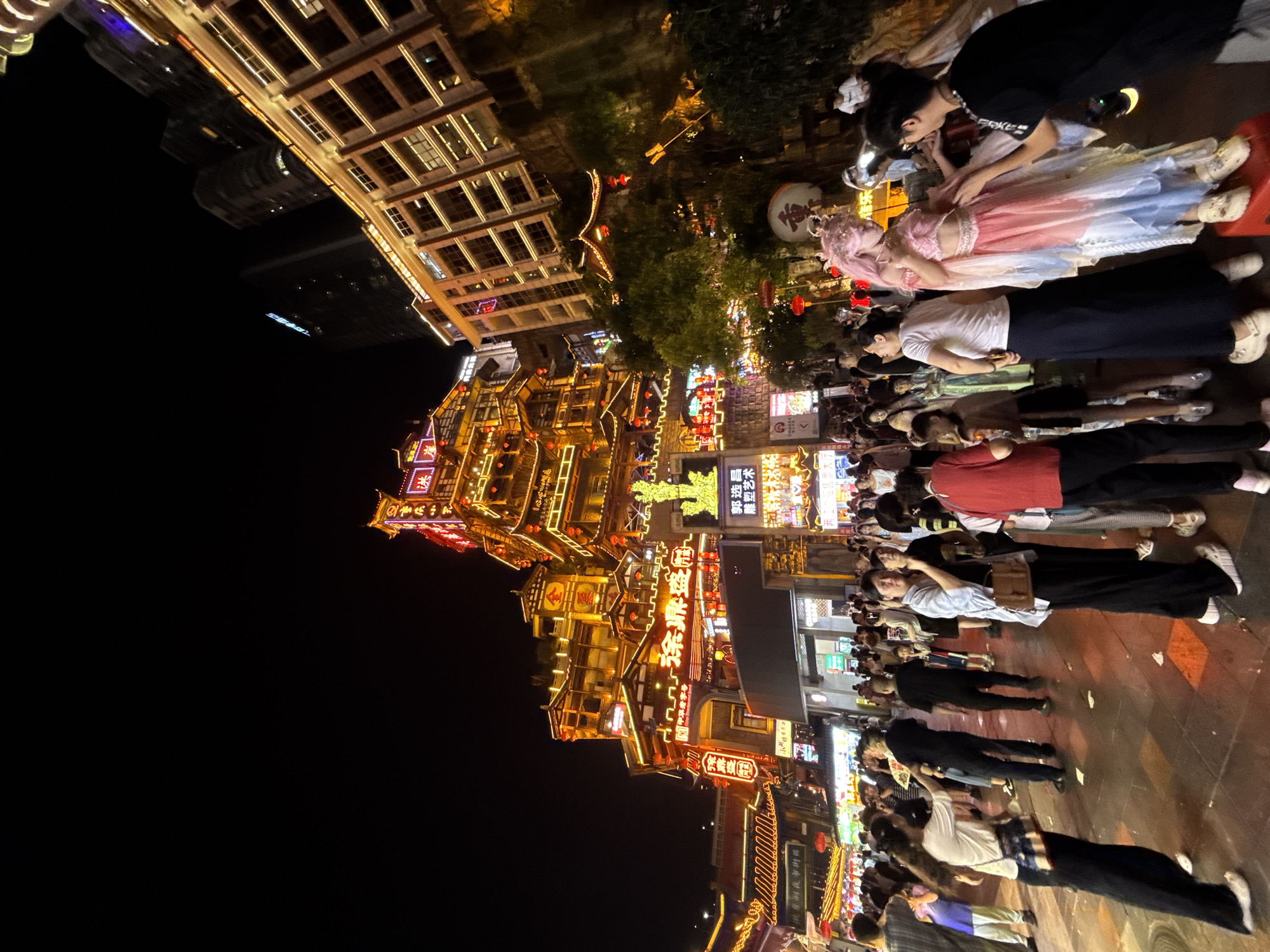 This is a nighttime, eye-level, wide-angle shot of a bustling street in Yuzhong District, China. The architecture is a prominent feature, with multi-story buildings illuminated by numerous bright lights, showcasing intricate details and traditional Chinese design elements. The main structure on the left is a tall, layered building adorned with red lanterns and glowing yellow and red signage. To the right, a modern high-rise building stands with its windows lit, contrasting with the traditional architecture.
The street is densely populated with people, suggesting a lively tourist area or a popular gathering spot. Many individuals are dressed in casual attire, with some wearing face masks. Several people are captured in motion, walking, standing, or interacting. In the foreground, a person in a pink and blue costume, possibly an entertainer or cosplayer, is posing on a red stool while interacting with another person. Another individual in a white t-shirt and black pants is standing next to them, facing towards the costumed person. The ground is paved with patterned tiles, and there are scattered pieces of litter. The overall atmosphere appears vibrant and energetic, characteristic of a busy urban night scene.