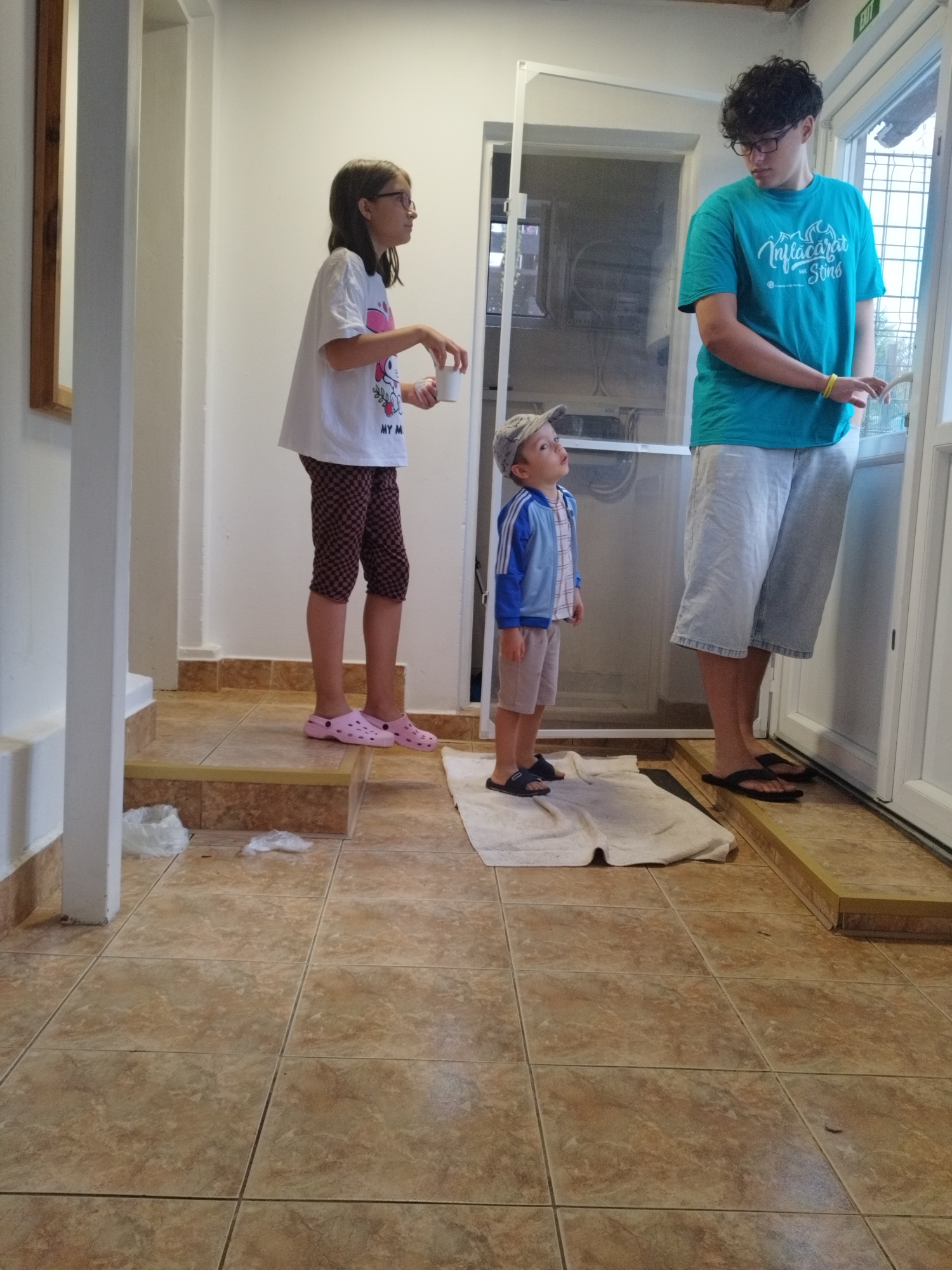 The image depicts an indoor scene featuring three individuals: a teenage boy, a pre-teen girl, and a young boy. The teenage boy, wearing glasses and a teal t-shirt, stands near a door, holding a white object. The pre-teen girl, with glasses and a white t-shirt, stands on a step holding a white cup. The young boy, wearing a blue jacket and a cap, looks upwards. The environment appears to be a hallway or entryway with tiled flooring and a wooden accent on the wall. The scene does not contain any identifiable landmarks or elements that would place it specifically in Bucharest, Romania.
