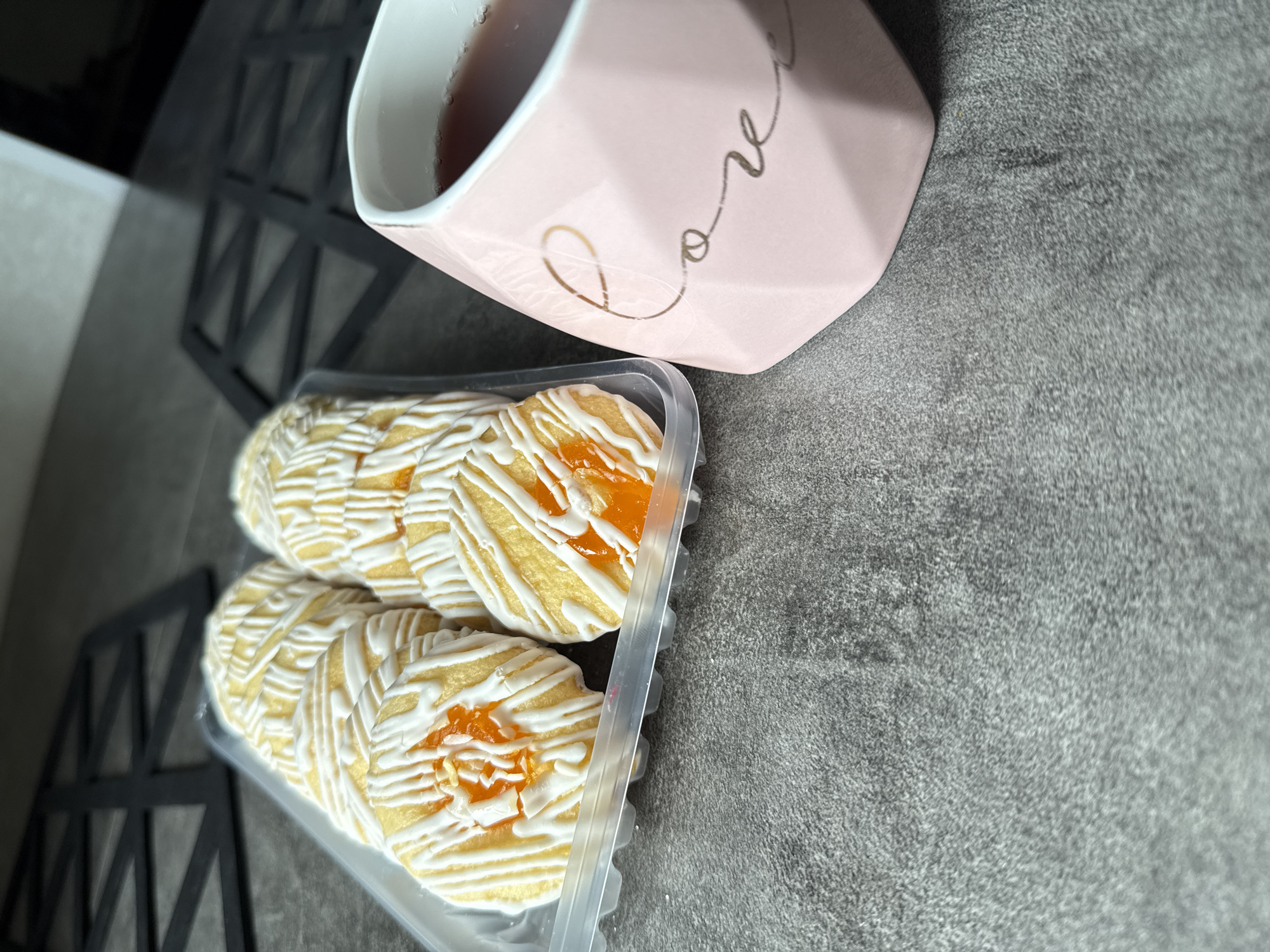 A close-up shot shows a pink mug with the word "love" written on it and a clear plastic container holding several round cookies. Each cookie is topped with a yellow filling and drizzled with white icing. The table has a gray, slightly textured surface. In the background on the left, a black, geometric patterned object is partially visible. There is no evidence suggesting the location is Petropavlovsk-Kamchatsky, Russia. The scene appears to be indoors. No people are visible.