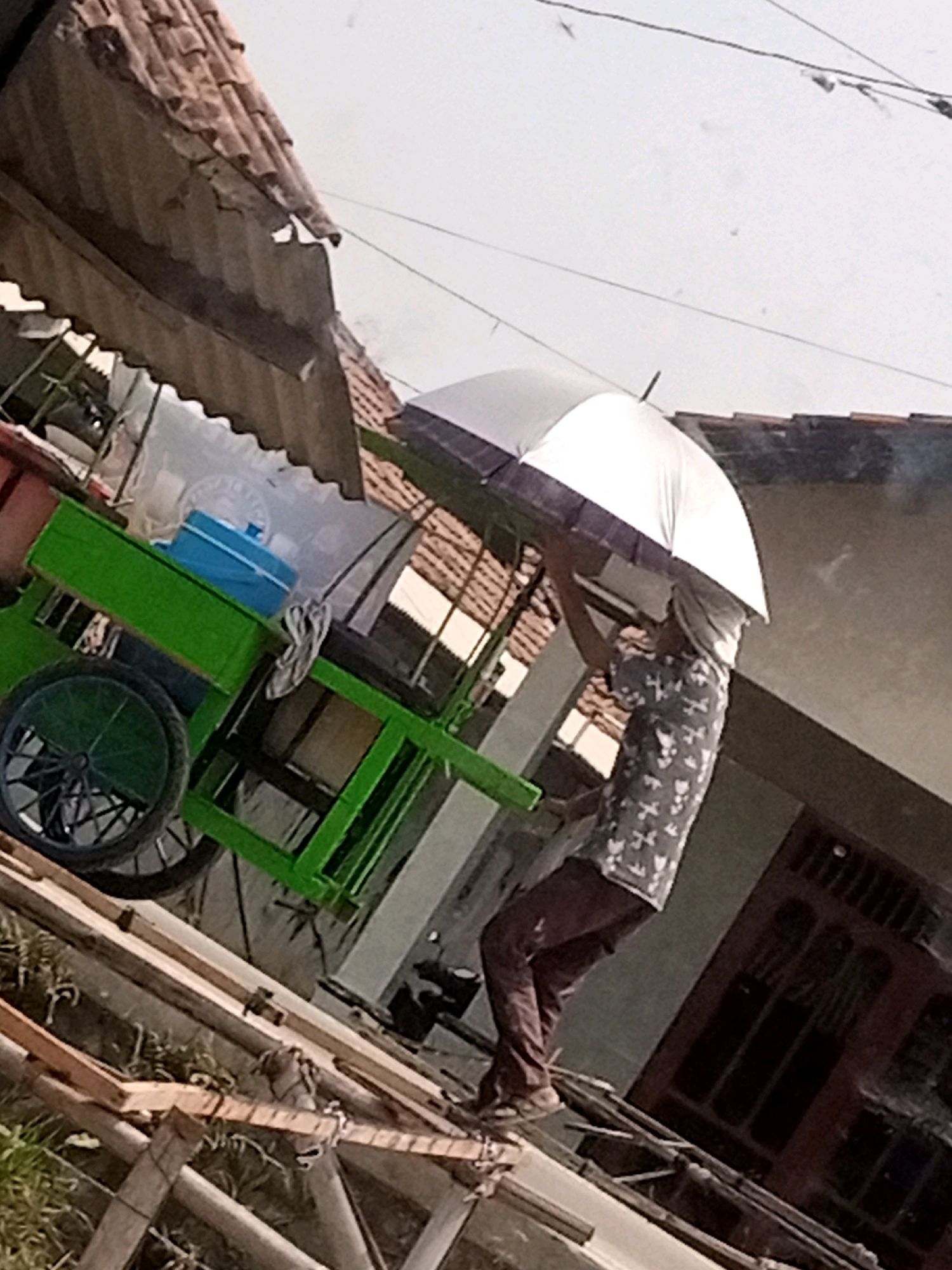 An individual stands on a wooden structure, holding a partially open umbrella above their head. The person is wearing a patterned shirt and dark pants. To the left of the individual is a green cart with a wheel. The scene appears to be outdoors, with structures resembling houses and roofing in the background, under a bright sky. The image is angled, suggesting it was taken from a low perspective. The location is identified as Boyoteluk, Indonesia.