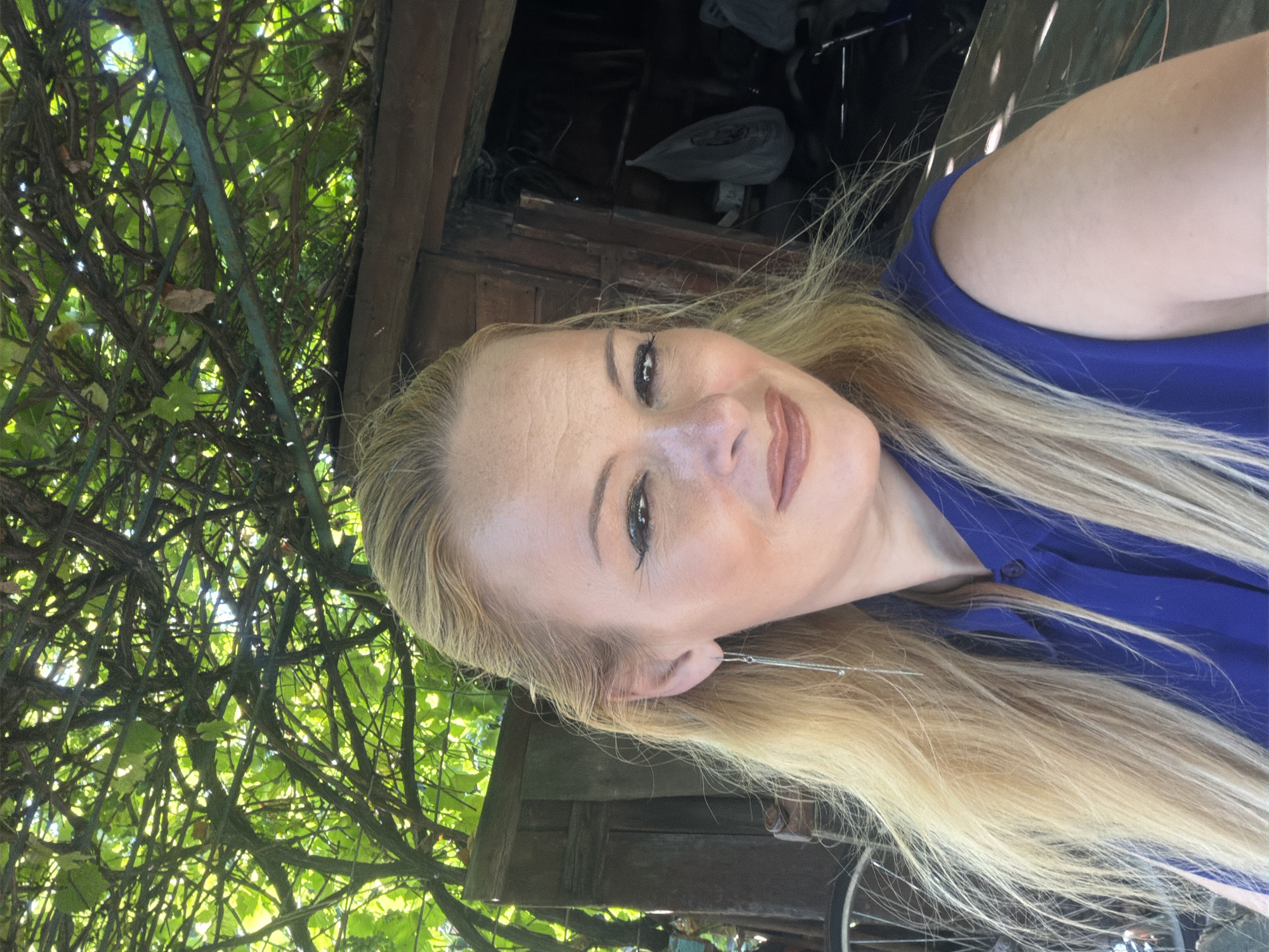 An outdoor, waist-up, eye-level medium shot captures a fair-skinned woman with blonde hair and light makeup posing for a selfie under a canopy of green vines. The woman is wearing a royal blue, collared, sleeveless shirt, and a long, delicate silver earring hangs from her left ear. She is looking directly at the camera with a slight smile and her mouth slightly open. Behind her, to her left, is the edge of a bicycle wheel, and to her right is a weathered wooden structure with an open doorway, revealing dark interior space and some shadowy objects. The overall lighting suggests a sunny day, with dappled sunlight filtering through the leaves overhead. The location is not discernable within the city of Unknown, Unknown.