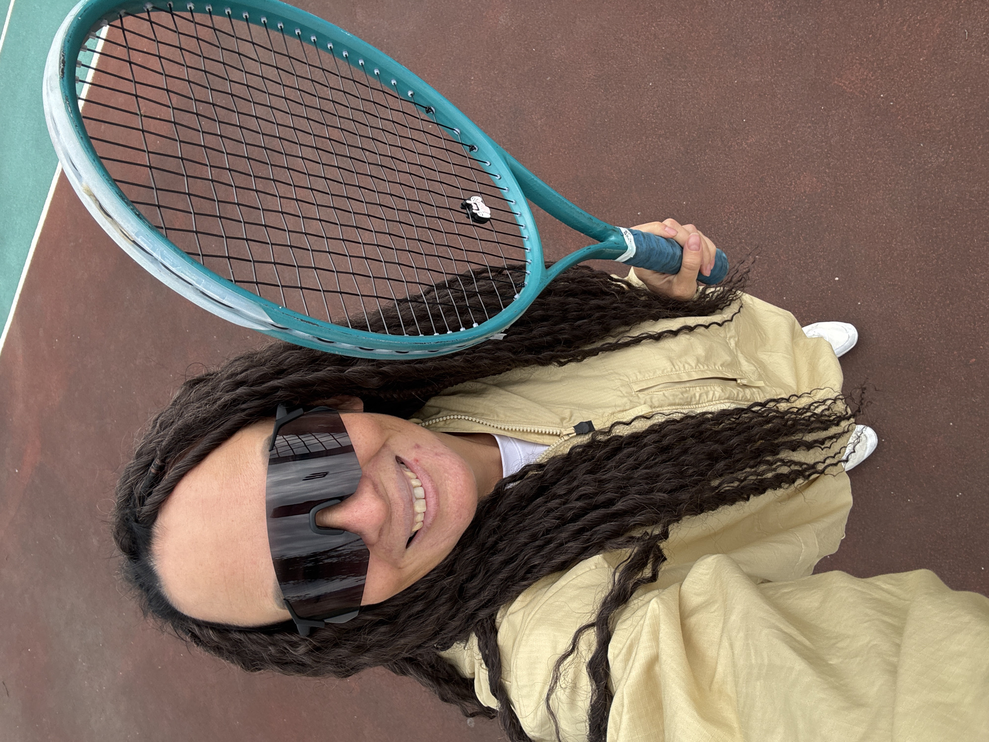 A high-angle selfie captures a person smiling brightly on a tennis court in Chaikovsky, Russia. The individual, seen from the chest up, has long, dark, textured hair (appearing as braids or micro-braids) and is looking up directly at the camera. They are wearing large, dark, sporty wrap-around sunglasses that obscure their eyes, reflecting the surroundings. A subtle logo, possibly a stylized 'B' or '3', is visible on the sunglasses. The person is dressed in a light-beige or khaki lightweight zippered jacket over a white top.
In their right hand, they hold a teal-colored tennis racket with a blue grip, extending it upwards and slightly to the left. The black strings of the racket feature a small white string dampener shaped like a miniature flip-flop or sandal, with faint text "ST" visible on it. The text "BLX" can be seen on the racket's blue grip.
The setting is an outdoor tennis court with a reddish-brown playing surface. A distinct green section with white boundary lines is visible in the upper left corner of the frame, indicating the court's layout. The bright lighting suggests it is daytime. A pair of white sneakers is partially visible near the bottom right. The person's broad smile conveys happiness and a cheerful mood.