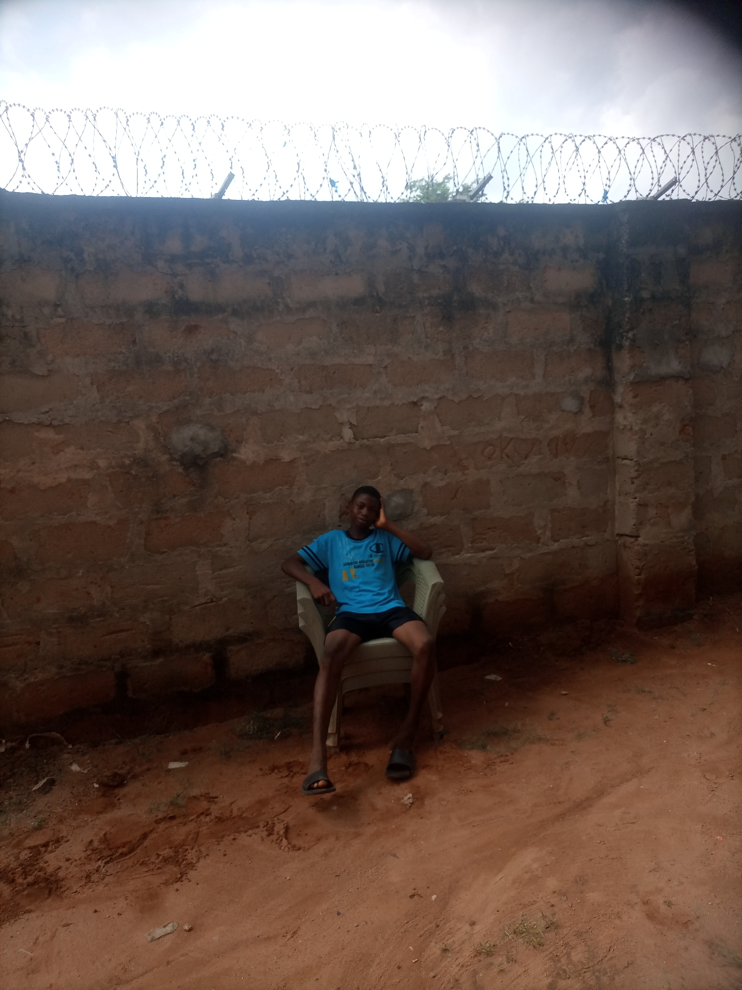 A young Black male, appearing to be in his late teens or early twenties, is seated in a white plastic chair against a weathered, rough-hewn brick wall. He is wearing a blue t-shirt with yellow lettering and black shorts, and dark flip-flops. His left arm is resting on the back of the chair, with his hand touching his head, and his right arm is resting on his leg. The background wall is made of unpainted, uneven bricks, some appearing chipped and discolored. A coiled barbed wire fence runs along the top of the wall. The ground in front of the wall is dry, reddish-brown earth with scattered debris. The sky above is overcast. The image appears to have been taken outdoors in a semi-urban or rural area, possibly Nkpor, Nigeria, given the architectural style of the wall and the general environment.