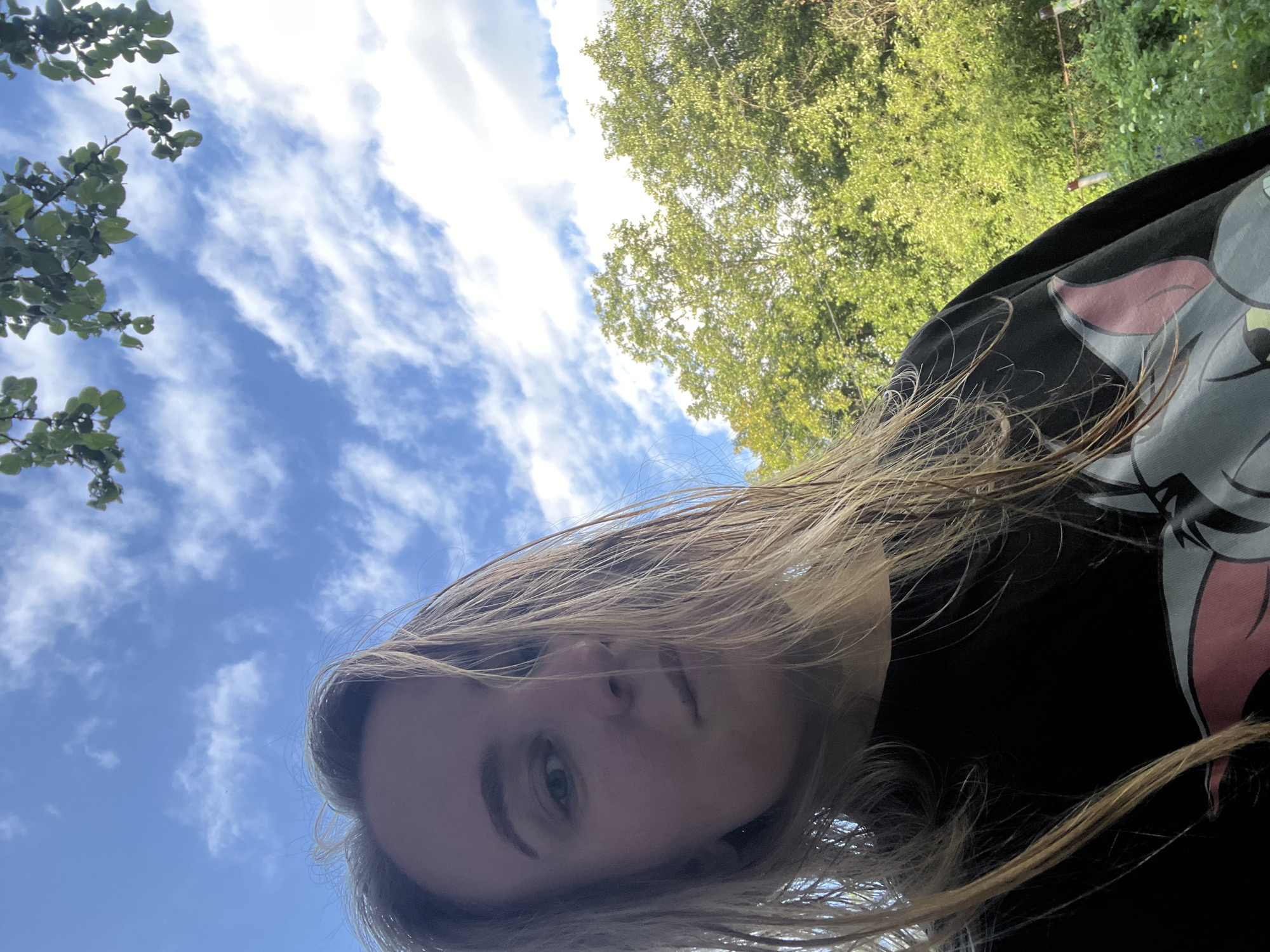 A selfie taken outdoors, likely on a cloudy day, shows a young woman with blonde hair framing her face. She is wearing a black t-shirt with a cartoon character printed on it, partially visible. The background consists of lush green trees and a blue sky with scattered white clouds. The image does not provide specific geographical markers to pinpoint a location within Chaikovsky, Russia.