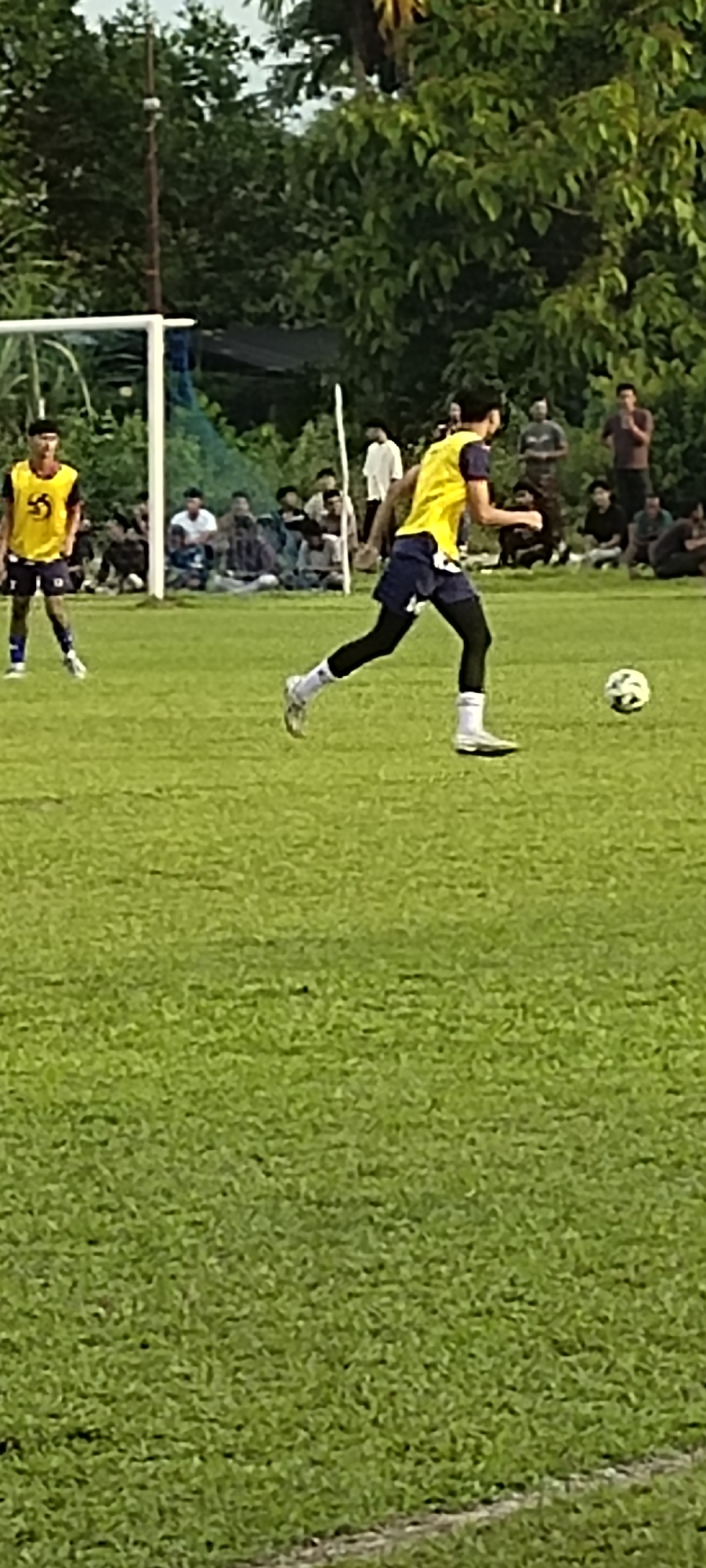 In Keudesimpangempat, Indonesia, a soccer match is underway on a grassy field. The primary focus is on two young men in soccer uniforms. One player, wearing a yellow jersey with a blue emblem and dark shorts with white socks, is in motion, dribbling a white soccer ball with his right foot. He is also wearing black compression pants underneath his shorts. The second player, also in a yellow jersey with an emblem and dark shorts, stands further back on the left side of the frame, observing the play. Behind the players, spectators are seated and standing, some obscured by trees. A soccer goal with a white frame and netting is visible on the far left, with trees and foliage forming the background. The scene captures a moment of active play during a soccer game.