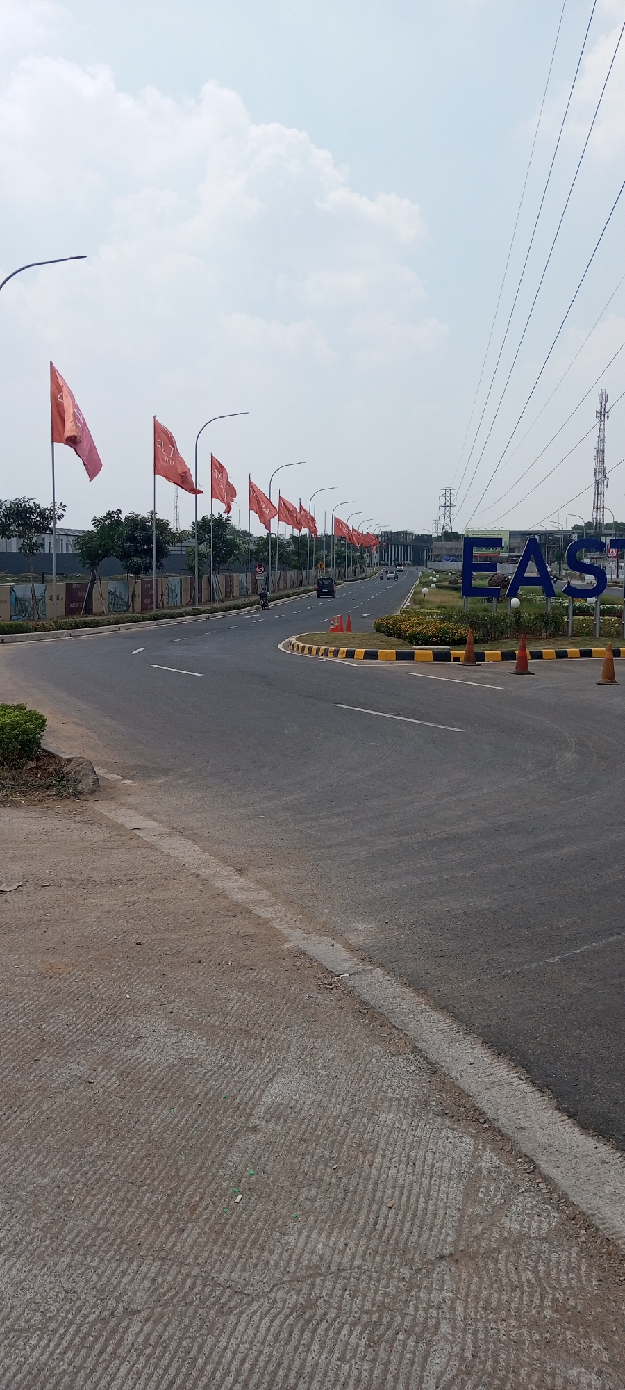 The image depicts an outdoor scene on a road in Rawa Banteng, Indonesia, likely a commercial or residential development. Red flags on poles line the left side of the road, a common sight in Indonesia during flag-related events or for promotional purposes. The road curves to the right, with a sidewalk made of textured concrete on the left and a grassy median with decorative plants and large blue letters spelling "EAST" on the right. Traffic cones are placed along the median's edge. In the background, there are buildings, trees, and a power transmission tower. The sky is overcast with clouds. There are a few vehicles on the road, including cars and a motorcycle. No people are clearly visible. The overall impression is one of a new development or an area undergoing construction.