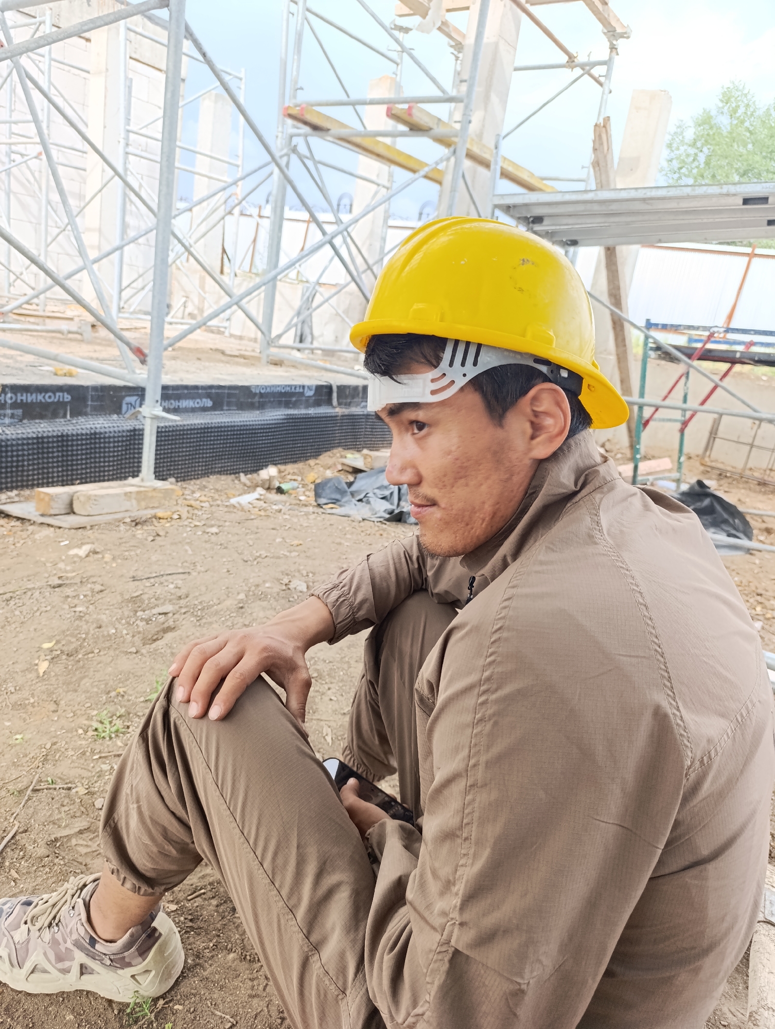 A medium shot shows a person wearing a yellow hard hat and a brown jumpsuit sitting on the ground at a construction site. The person is looking to their right and holding a mobile phone. The person's left hand is resting on their left knee. Their right leg is bent at the knee, and their right foot is resting on the ground. The person is wearing camouflage-patterned shoes. In the background, there is scaffolding, construction materials, and some trees. The ground is covered in dirt and gravel. The sky is overcast. The scene appears to be in an outdoor construction area.