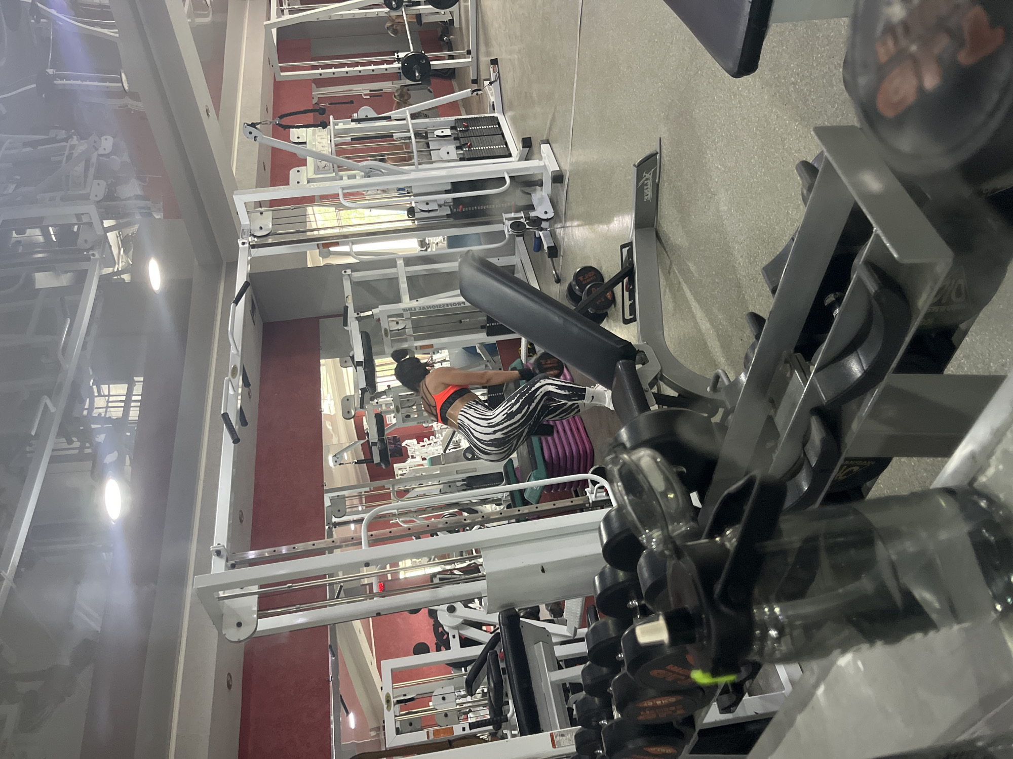 The image depicts the interior of a gym in Temirtau, Kazakhstan. In the foreground, a rack of dumbbells is visible, with a water bottle on a nearby surface. The central focus is a woman with dark hair, wearing an orange sports bra and black and white striped leggings, performing an exercise with weights on a bench. She is positioned in front of a multi-gym machine. The gym environment includes various weight machines, mirrors reflecting the ceiling lights and gym equipment, and a red-colored wall. The flooring appears to be a smooth, grey surface.