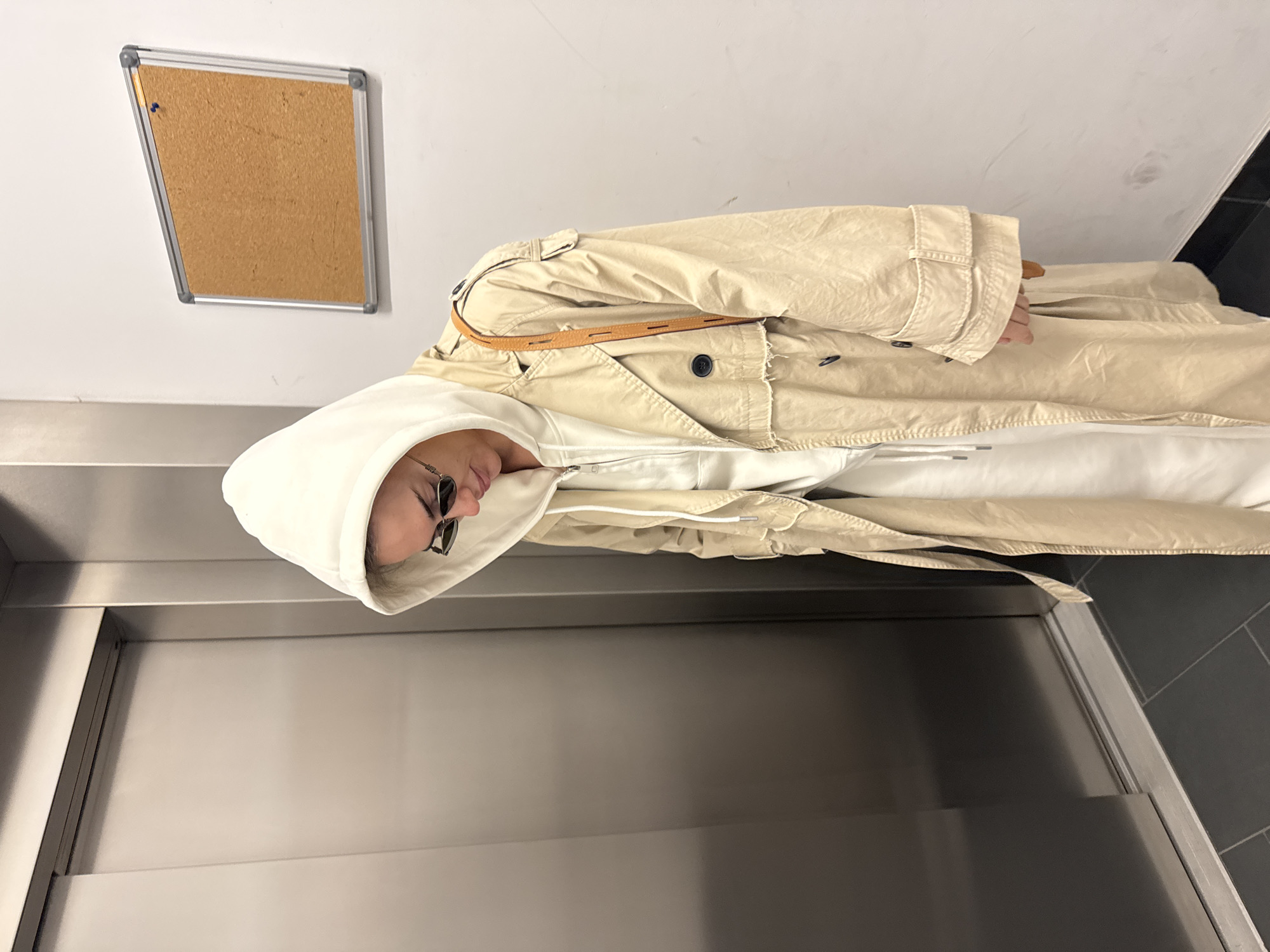 A person wearing a beige trench coat and a white hoodie is standing in front of an elevator. The person is wearing sunglasses. A corkboard is mounted on the wall behind them. The location is Warsaw, Poland.