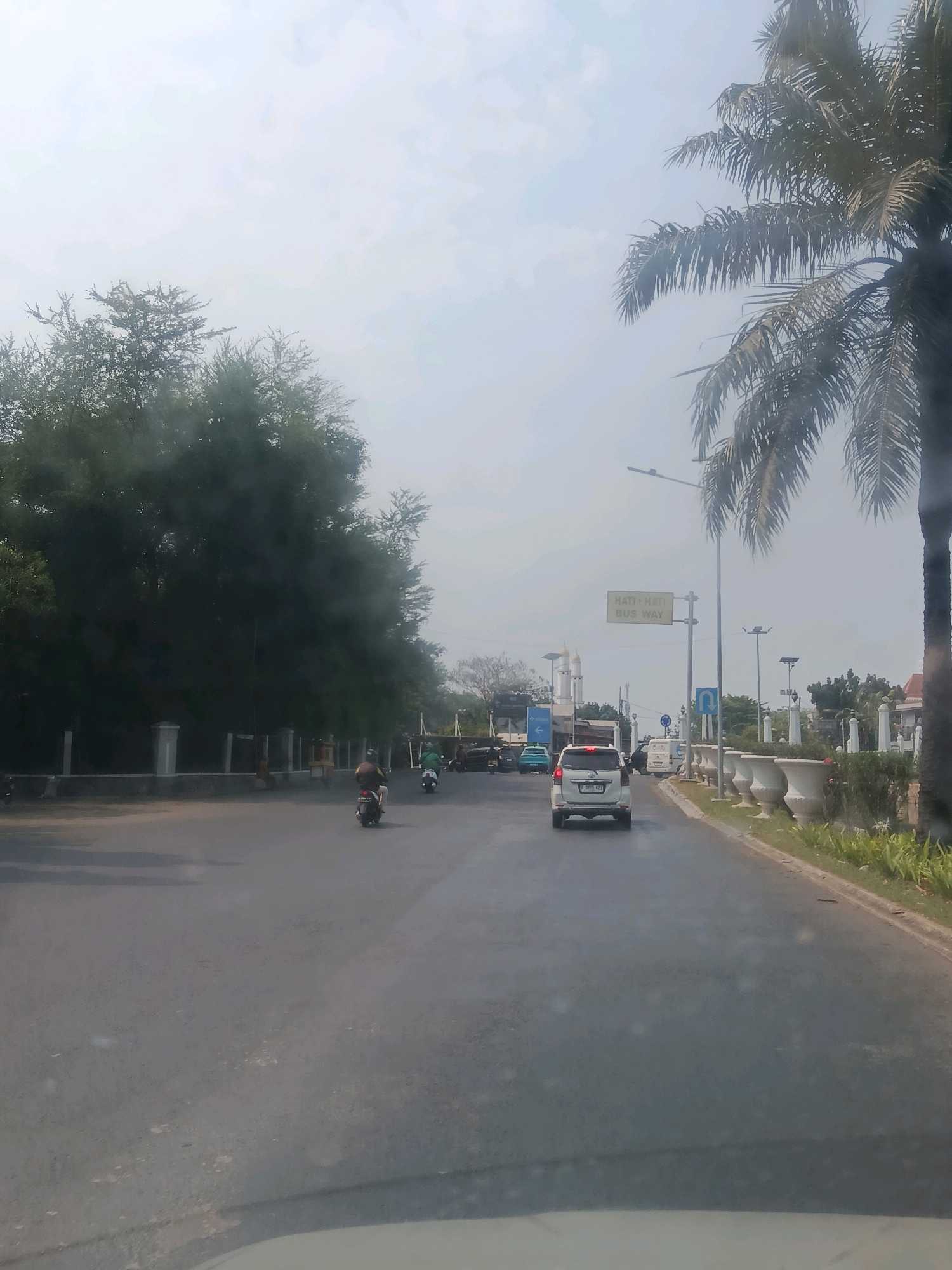 The image captures a view from a vehicle traveling on a road in Bekasi, Indonesia, on a hazy day. A white SUV is in the foreground, followed by a white minivan further ahead. Several motorcycles are also present on the road, with one rider in the middle ground visible from behind. To the left of the road, a line of white pillars and a low wall borders a dense area of green trees. On the right side of the road, a tall palm tree dominates the right edge of the frame, with a well-maintained sidewalk adorned with large decorative planters. A yellow sign with the text "HATI-HATI BUS WAY" is visible on a lamppost. In the background, white domed structures, possibly minarets, suggest the presence of a mosque. The overall environment appears to be an urban or suburban street with moderate traffic.