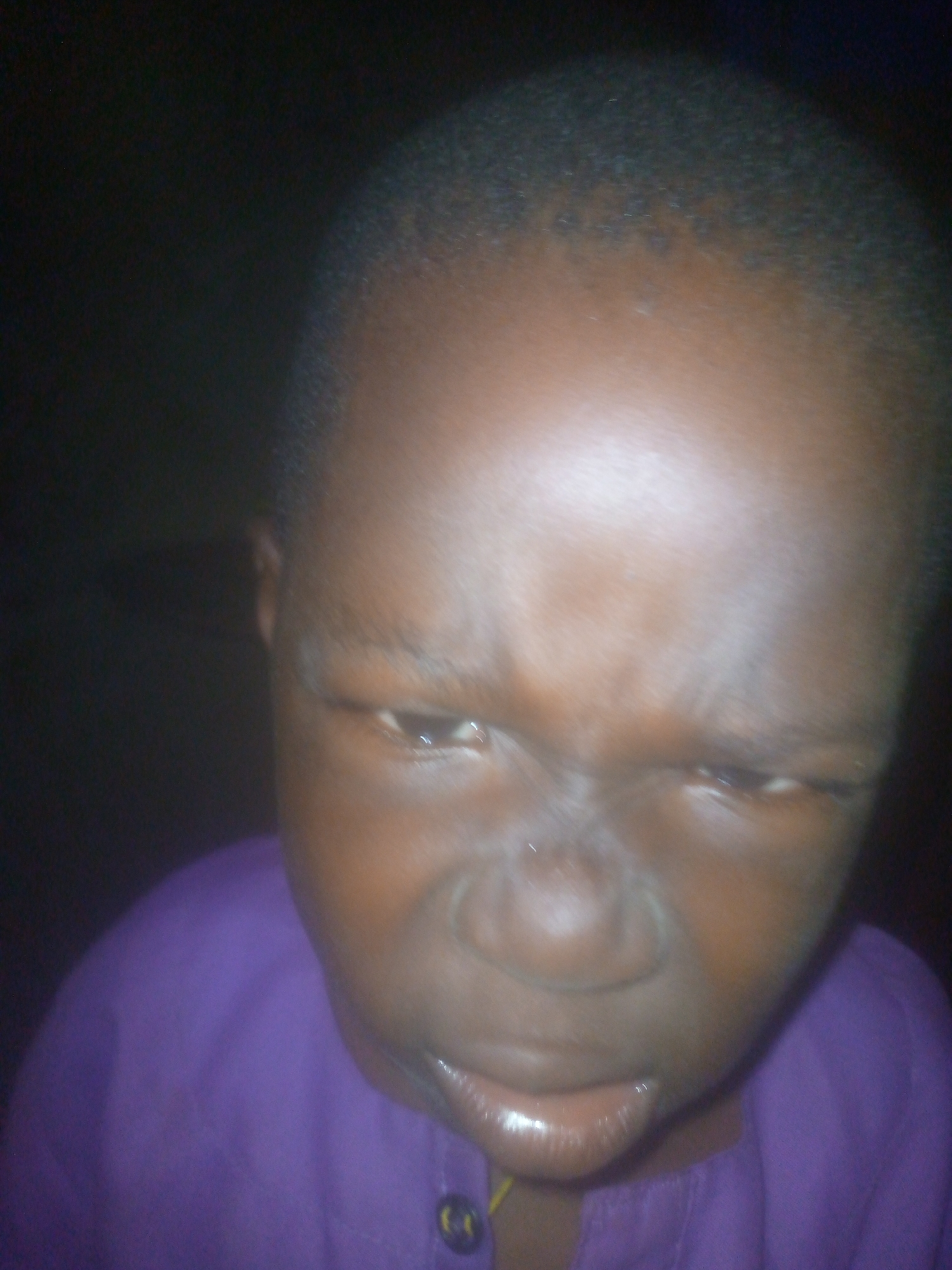 A close-up of a young Black boy with a furrowed brow. The image is dimly lit. He is wearing a purple shirt. The location is Unknown, Unknown.