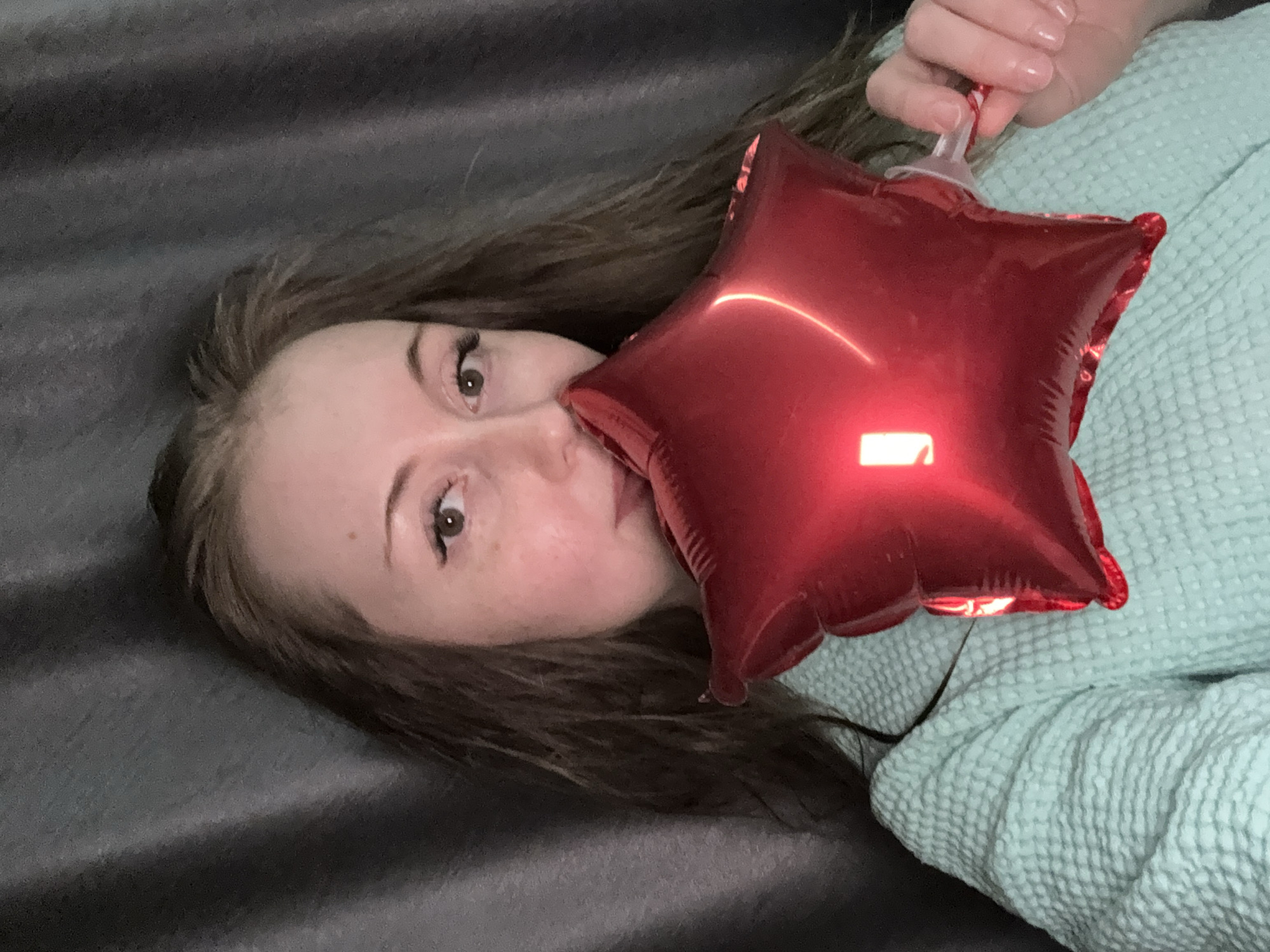 A young woman is depicted in a close-up, rotated portrait, lying on her side. Her long, dark brown hair is spread out behind her. She looks directly at the viewer with wide, brown eyes, featuring prominent eyelashes. Her fair skin shows subtle freckles. A bright, reflective red star-shaped balloon is held in front of her face, partially obscuring her mouth and nose. Her right hand, with neatly manicured nails, is visible holding the clear plastic stem of the balloon. She is wearing a light blue-green textured garment, possibly a robe or blanket, which covers her body. The background consists of a dark gray, textured fabric, likely a bedspread or blanket, suggesting an indoor setting such as a bedroom or living room. The lighting appears artificial, creating distinct highlights and reflections on the shiny surface of the balloon. No text is visible in the image. The overall impression is one of a playful or relaxed moment.